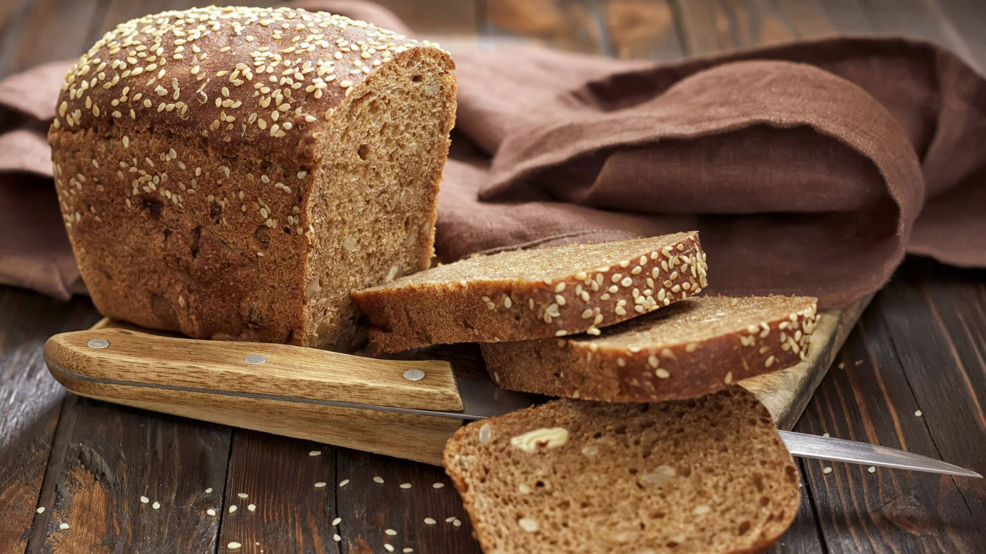 Bread 桌布 壁紙 — 新鮮出爐的全麥麵包撒上芝麻，切片放在木板上，旁邊有刀和棕色布。