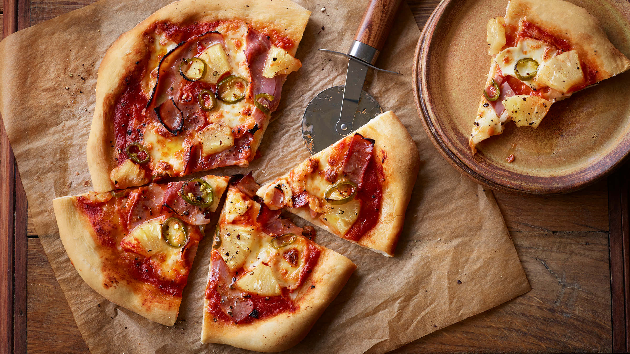 Pizza 桌布 壁紙 — 新鮮出爐的夏威夷披薩，有火腿、鳳梨和墨西哥辣椒，在烘焙紙上切片，旁邊有個木盤。