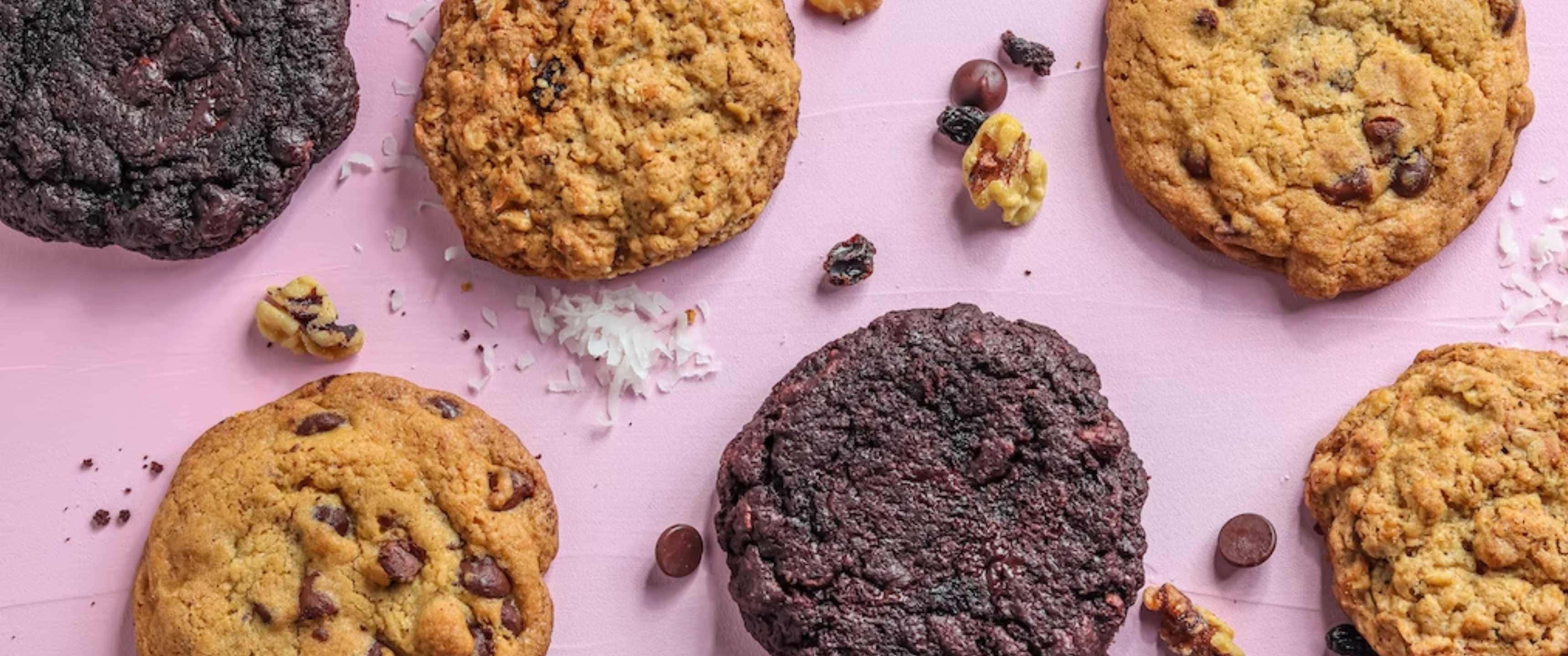 Cookies 桌布 壁紙 — 新鮮出爐的巧克力碎和燕麥餅乾散落在柔軟的粉紅色表面上，周圍還有散落的堅果和椰子片。