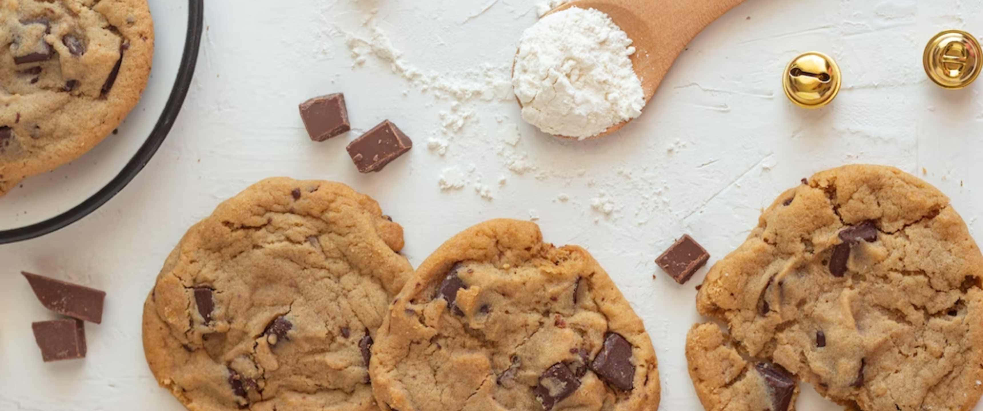 Cookies 桌布 壁紙 — 新鮮出爐的巧克力餅乾散落在白色表面上，伴有巧克力塊、麵粉和金色鈴鐺。