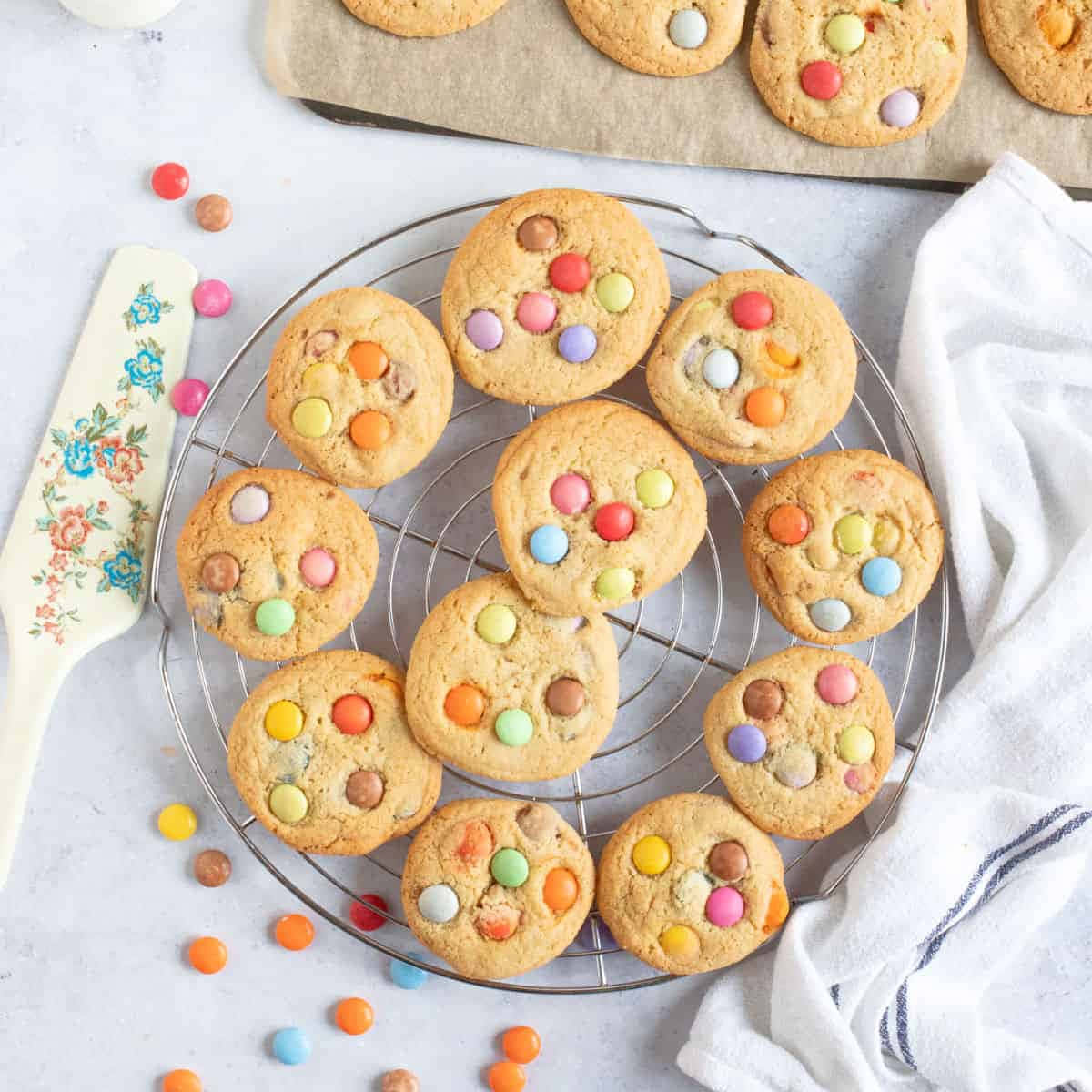 Cookie 桌布 壁紙 — 新鮮出爐的彩色M&M餅乾在金屬架上冷卻，旁邊有花卉圖案刮刀和白色毛巾。