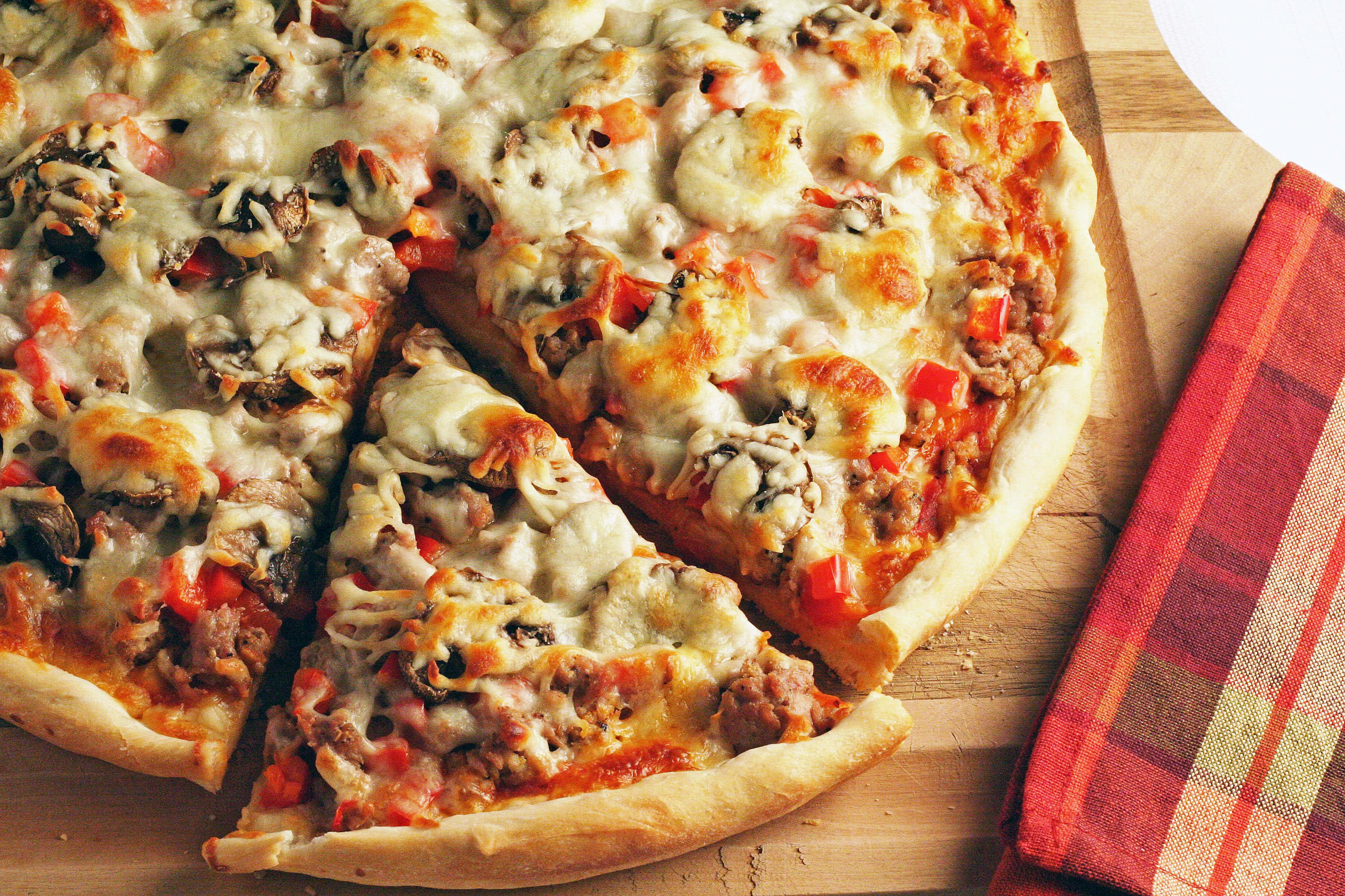 Pizza 桌布 壁紙 — 新鮮出爐的披薩，上面有融化的起司、蘑菇和紅椒，切片放在木板上，旁邊有一塊格子餐巾。
