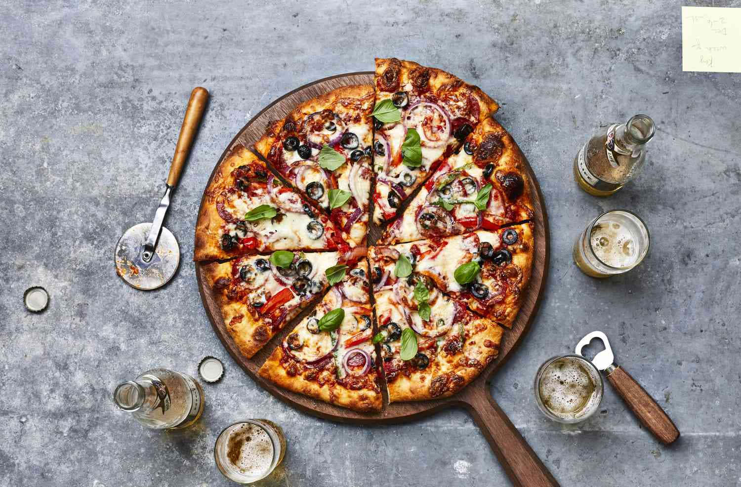 Pizza 桌布 壁紙 — 新鮮出爐的披薩，上面鋪有橄欖、洋蔥和羅勒，放在木板上，周圍有啤酒瓶和玻璃杯，置於灰色石面上。