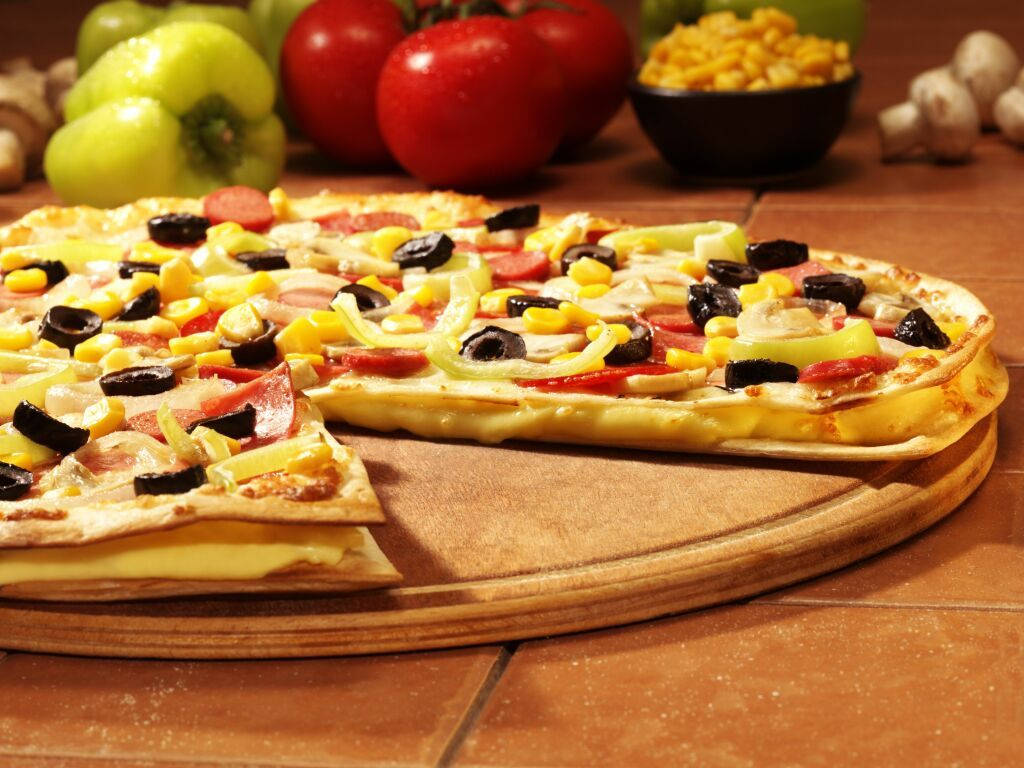 Dominos Pizza 桌布 壁紙 — 新鮮出爐的披薩，上面鋪有橄欖、甜椒、玉米和火腿，放在木板上，背景有食材。