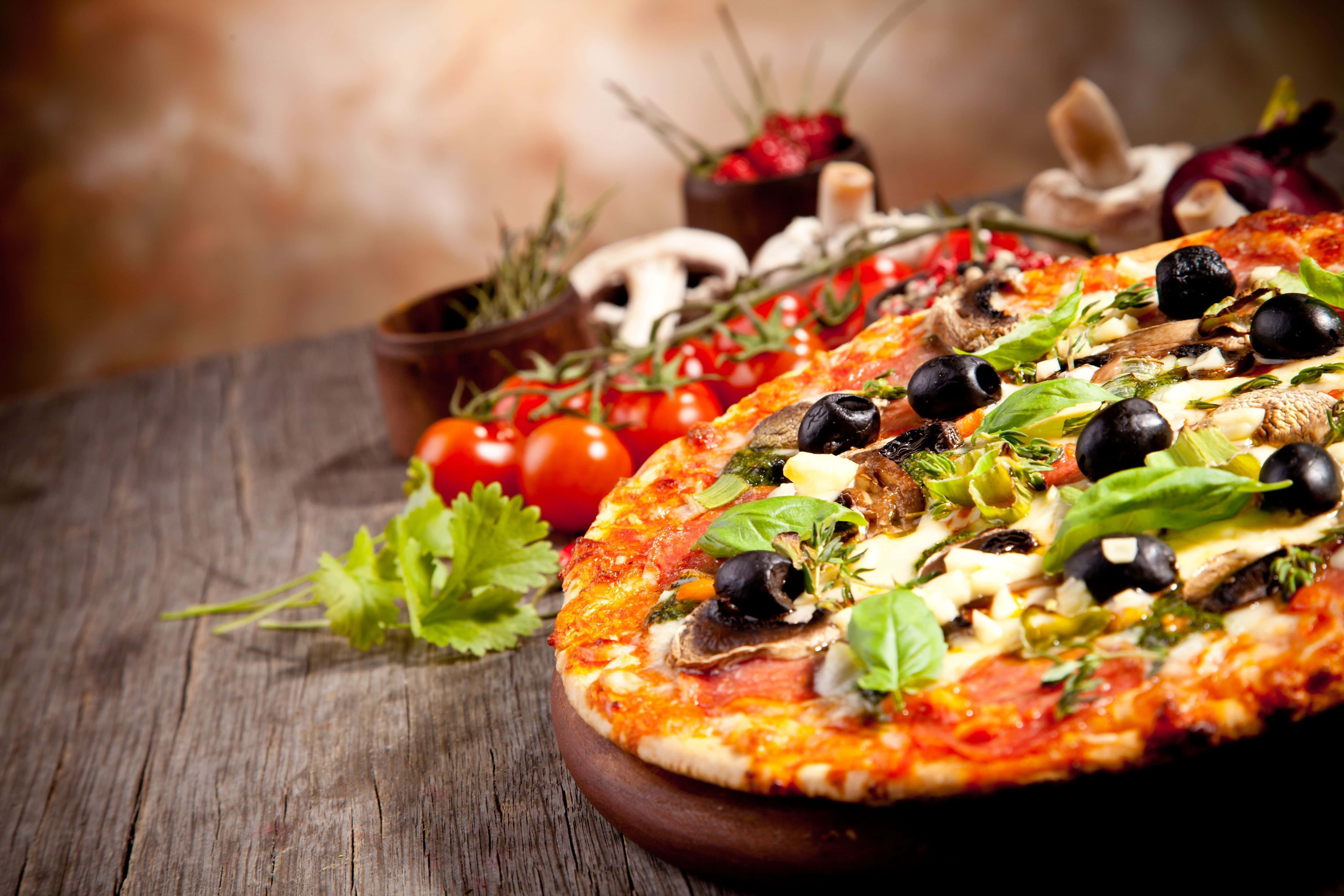 Pizza 桌布 壁紙 — 新鮮出爐的披薩，上面鋪有橄欖、蘑菇和羅勒，放在木板上，周圍環繞著鮮豔的櫻桃番茄和新鮮香草。