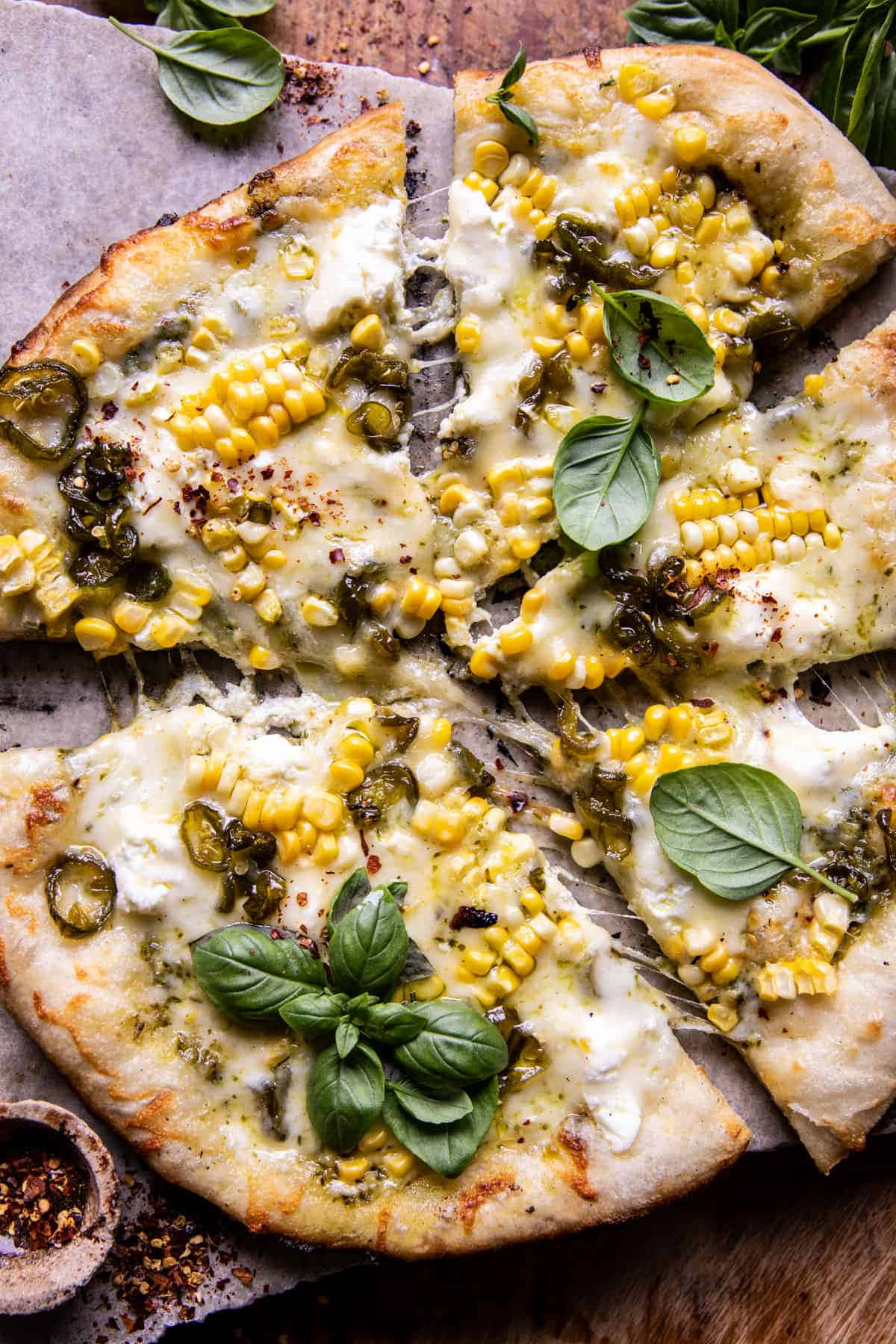 Pizza 桌布 壁紙 — 新鮮出爐的披薩，上面鋪滿玉米墨西哥辣椒香蒜醬和羅勒，切片放在烘焙紙上，旁邊有一小碗紅辣椒片。