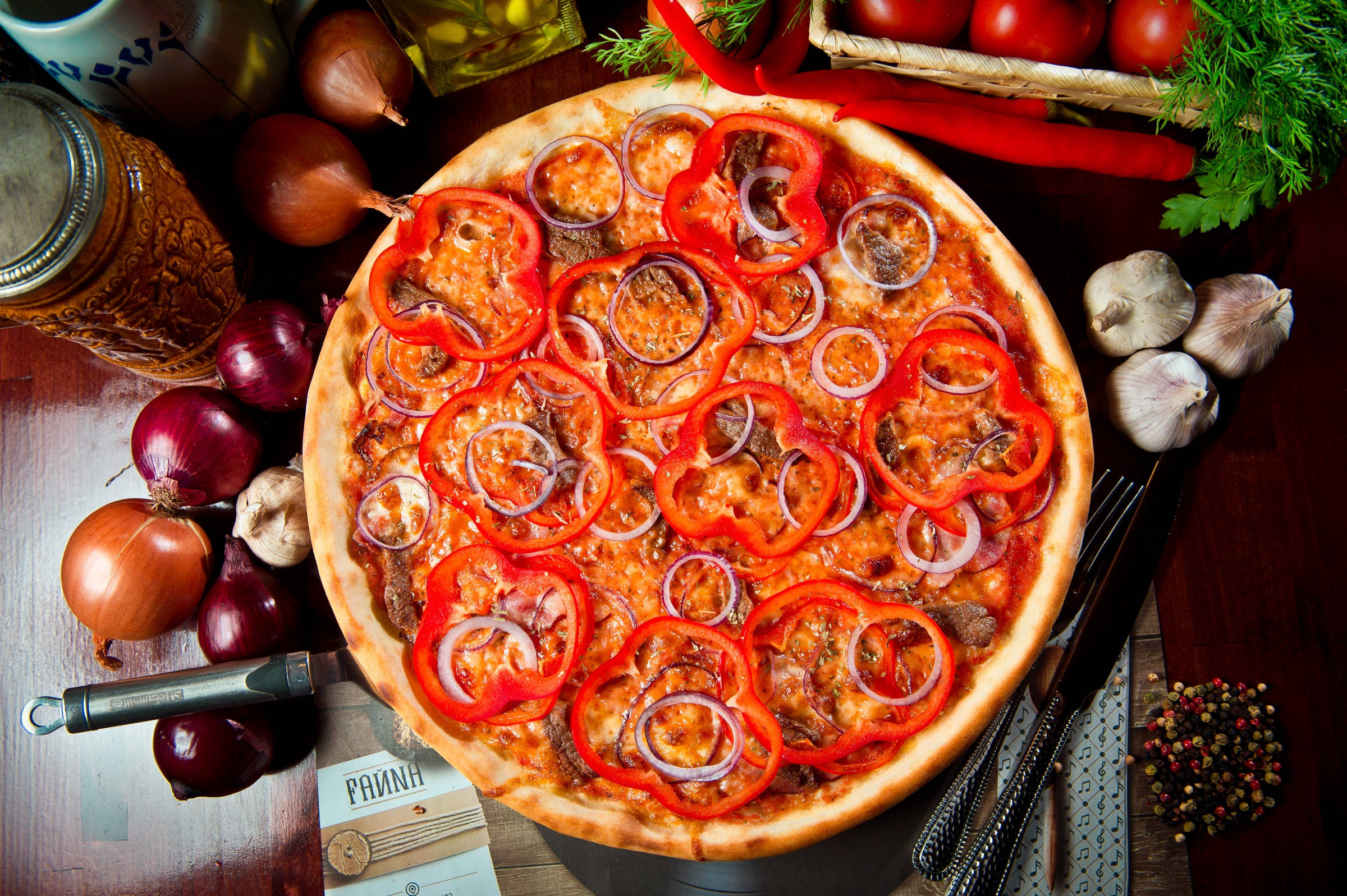 Pizza 桌布 壁紙 — 新鮮出爐的披薩，上面鋪滿紅椒和洋蔥，放在木桌上，周圍有蒜頭、番茄和香草。