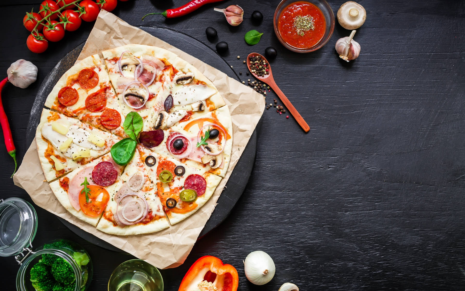 Pizza 桌布 壁紙 — 新鮮出爐的披薩，上面鋪滿義大利辣香腸、橄欖、洋蔥和羅勒，放在烘焙紙上，周圍是深色鄉村風格表面上的鮮豔食材。
