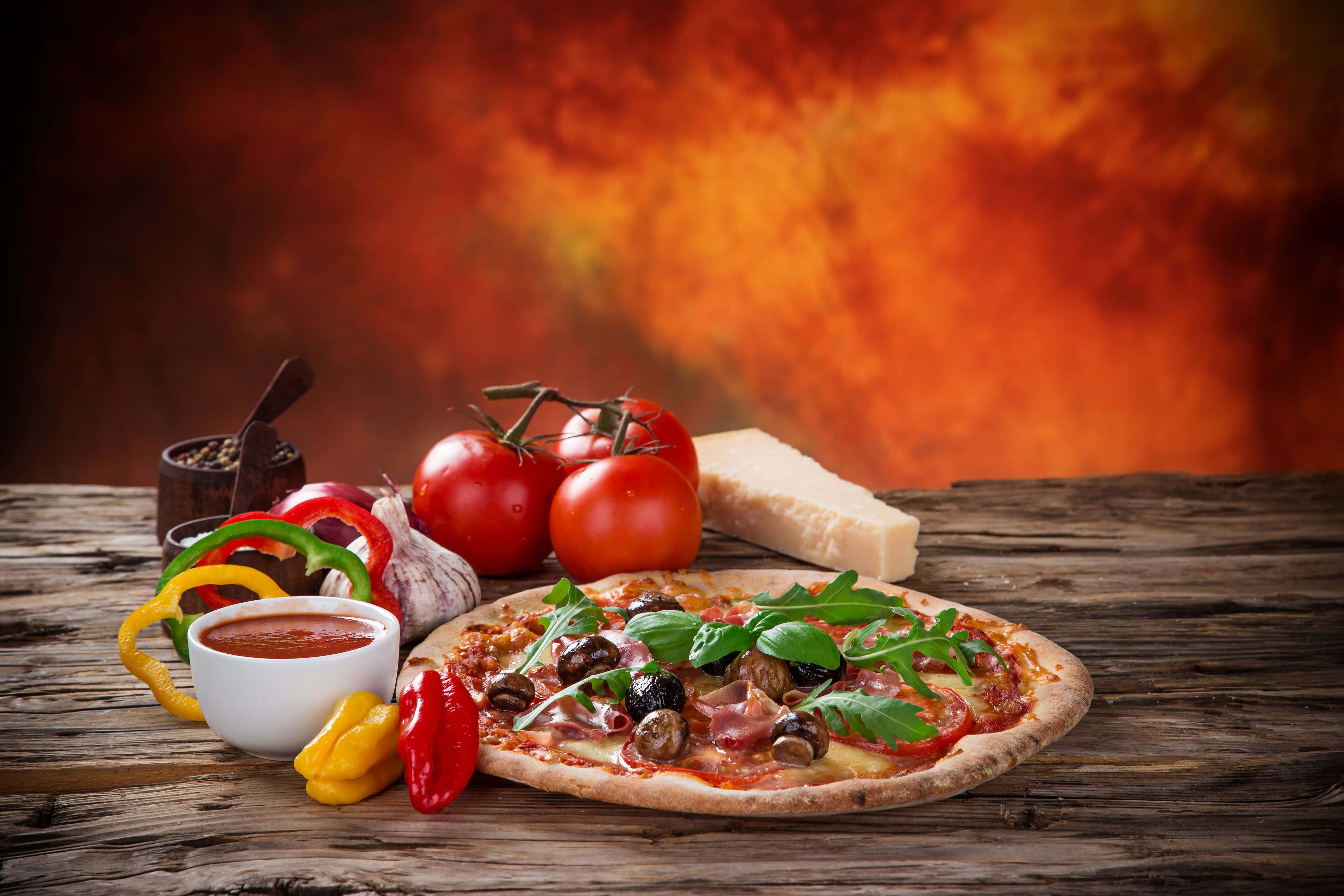 Pizza 桌布 壁紙 — 新鮮出爐的披薩，上面鋪滿蘑菇、芝麻葉和火腿，放在質樸的木桌上，周圍是色彩繽紛的蔬菜和起司，背景是火熱的橙色。