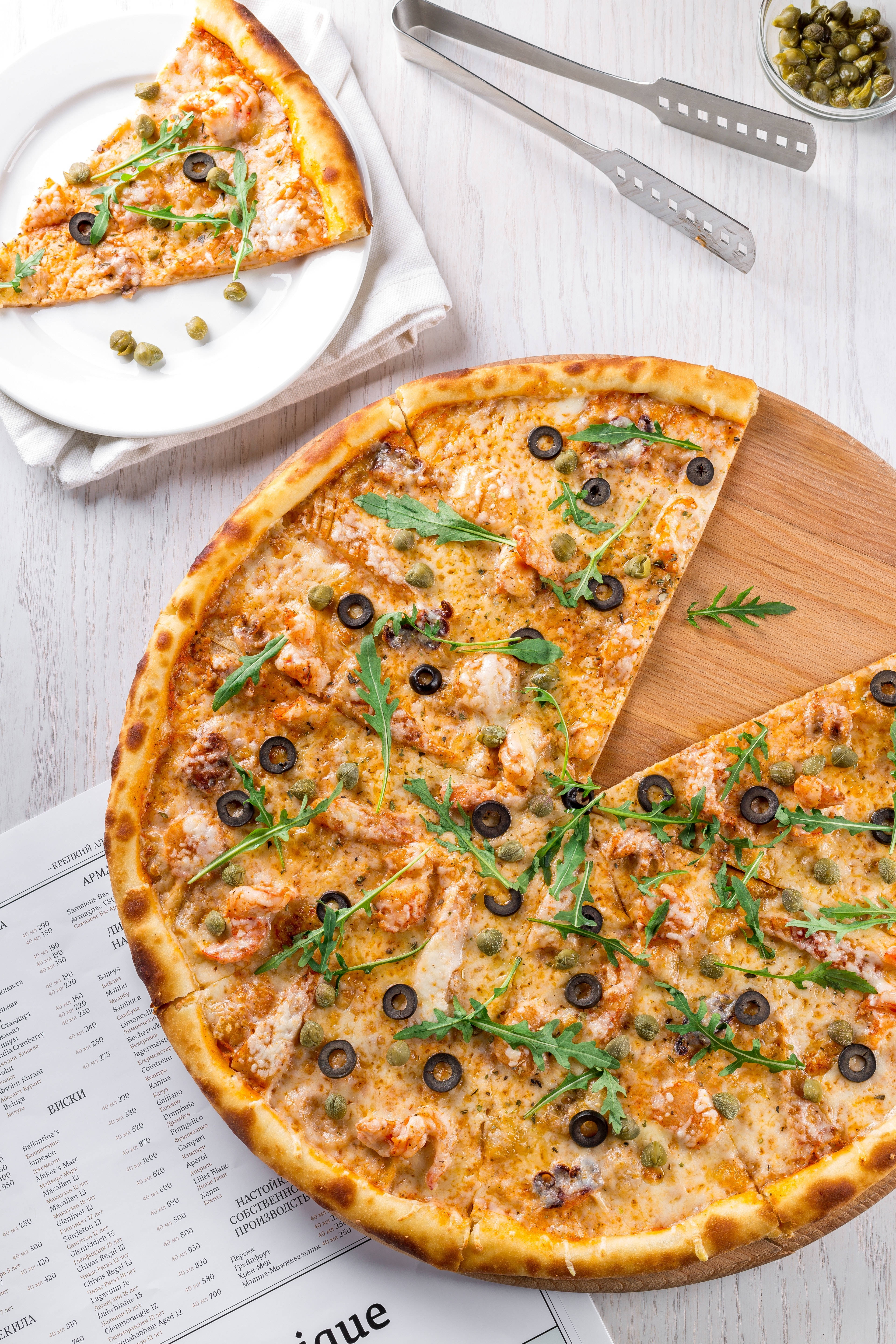 Dominos Pizza 桌布 壁紙 — 新鮮出爐的披薩，上面鋪滿蝦、芝麻葉和橄欖，放在木板上，旁邊有一片放在盤子上。