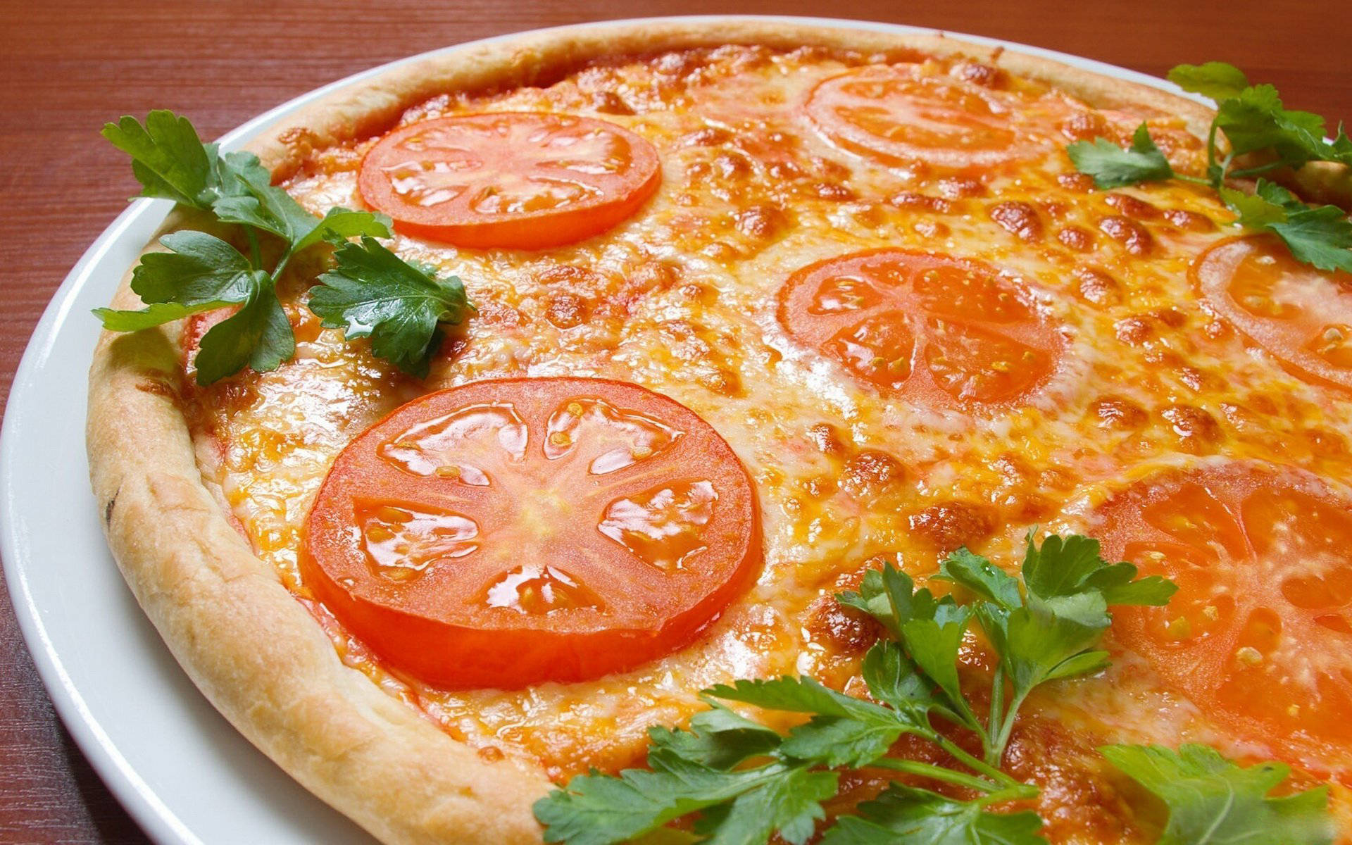 Pizza 桌布 壁紙 — 新鮮出爐的披薩，上面鋪滿融化起司和鮮紅色番茄片，並以綠色巴西里點綴，放在白色盤子上。