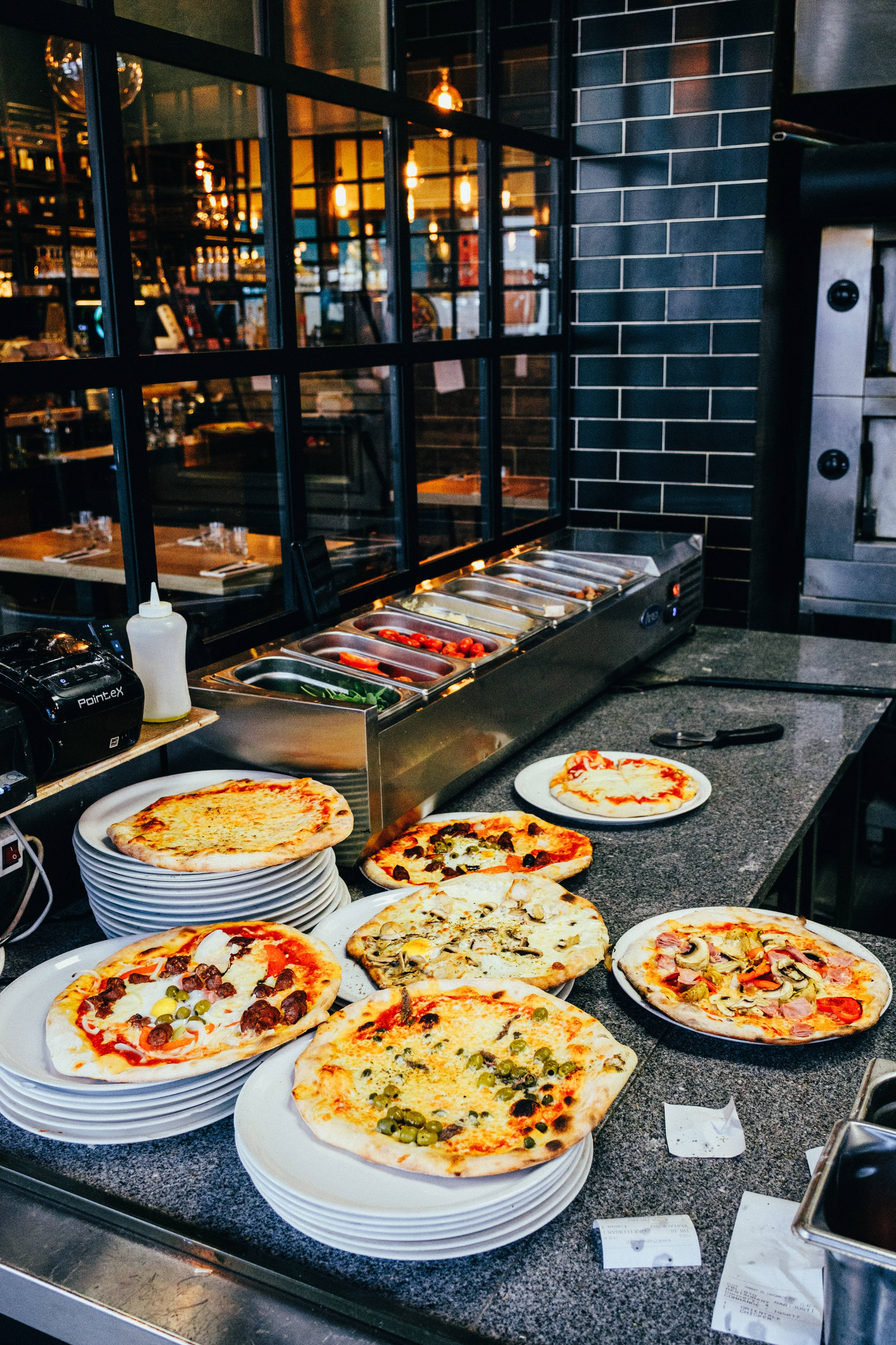 Dominos Pizza 桌布 壁紙 — 新鮮出爐的披薩配上色彩繽紛的配料，放在盤子上，旁邊是現代披薩店的不鏽鋼自助餐檯。