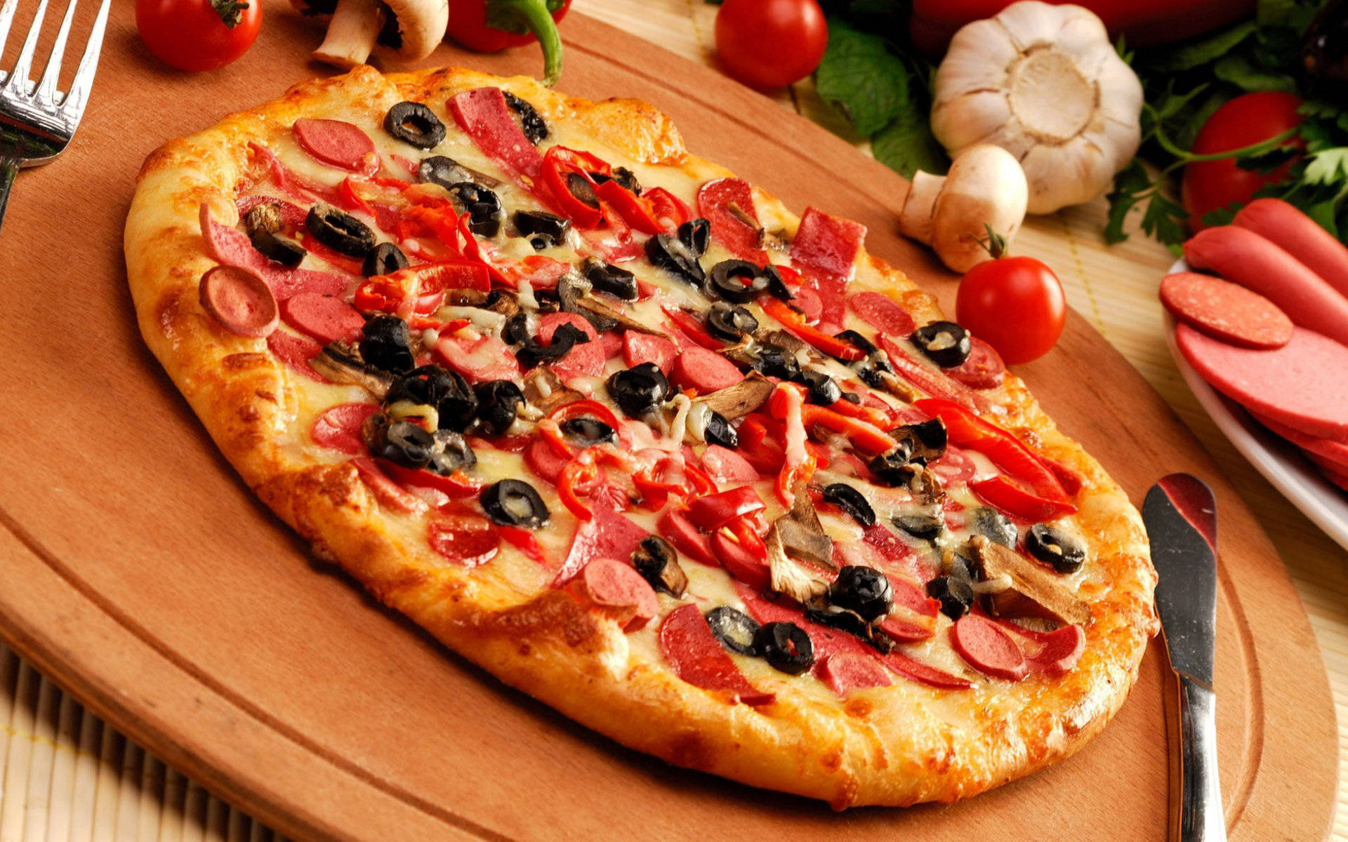 Pizza 桌布 壁紙 — 新鮮出爐的橢圓形披薩，上面鋪滿義大利辣香腸、橄欖、甜椒和蘑菇，放在木板上，周圍環繞著新鮮食材。