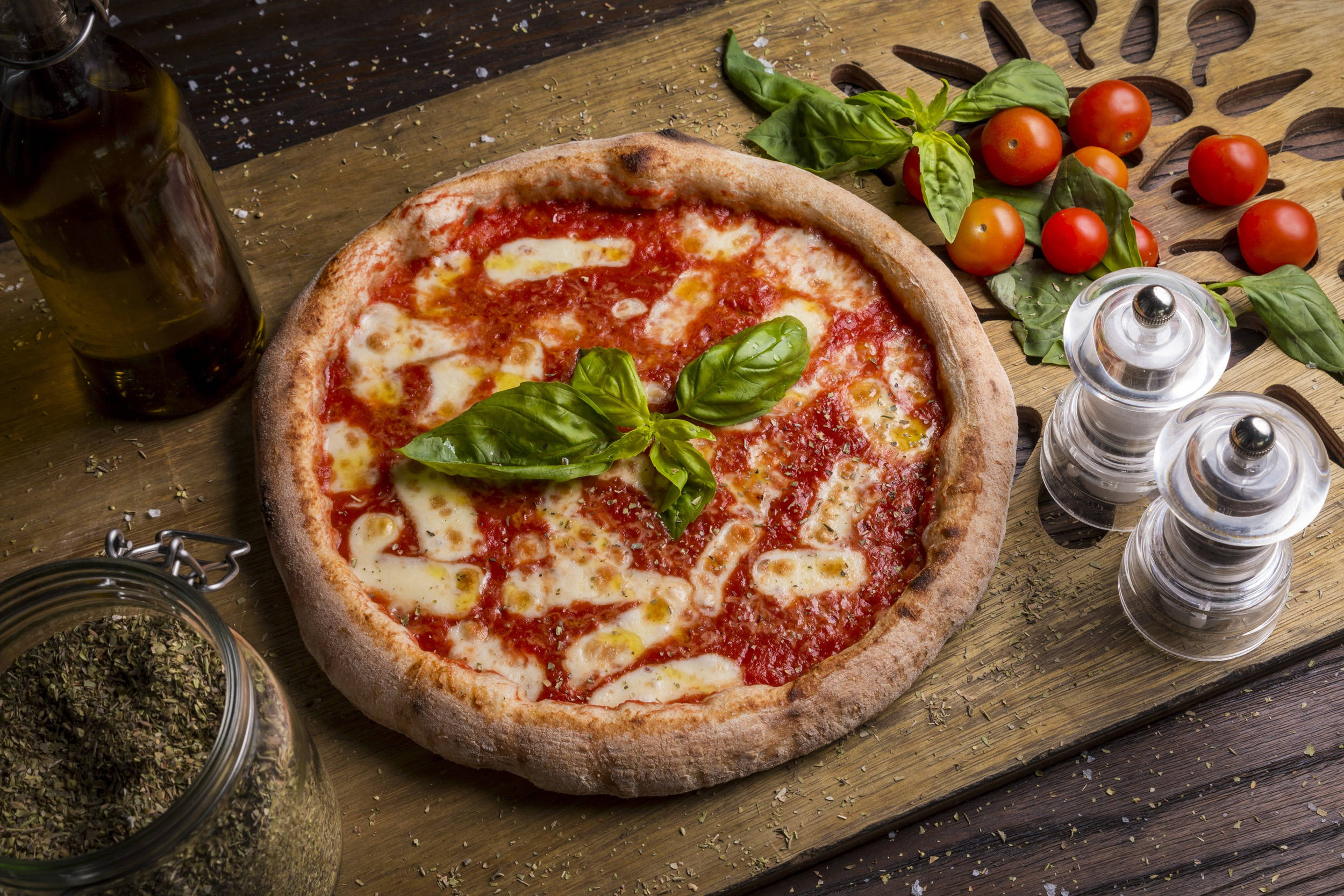 Pizza 桌布 壁紙 — 新鮮出爐的瑪格麗特披薩，上面有融化的莫扎瑞拉起司和羅勒葉，放在木板上，周圍環繞著櫻桃番茄、橄欖油和調味料。