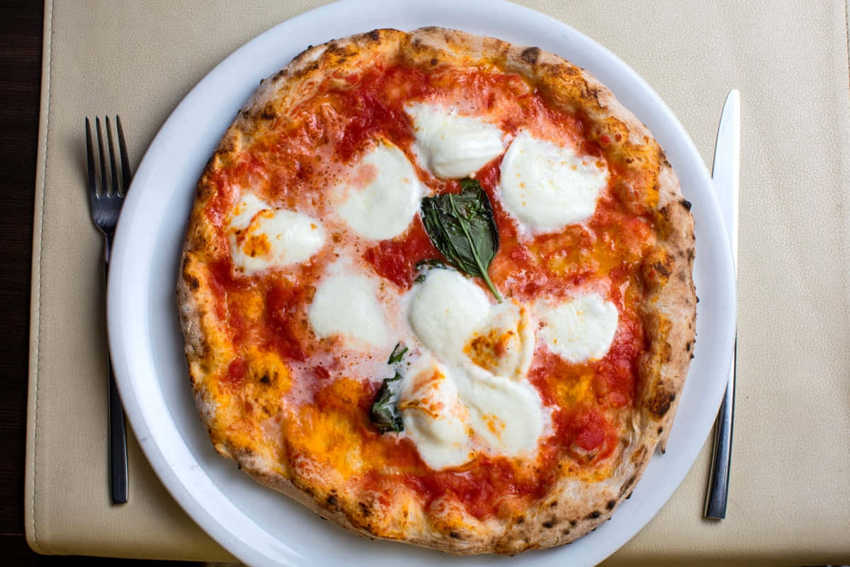 Pizza 桌布 壁紙 — 新鮮出爐的瑪格麗特披薩，配上融化的莫薩里拉起司、羅勒和番茄醬，放在白色盤子上，旁邊有叉子和刀子。