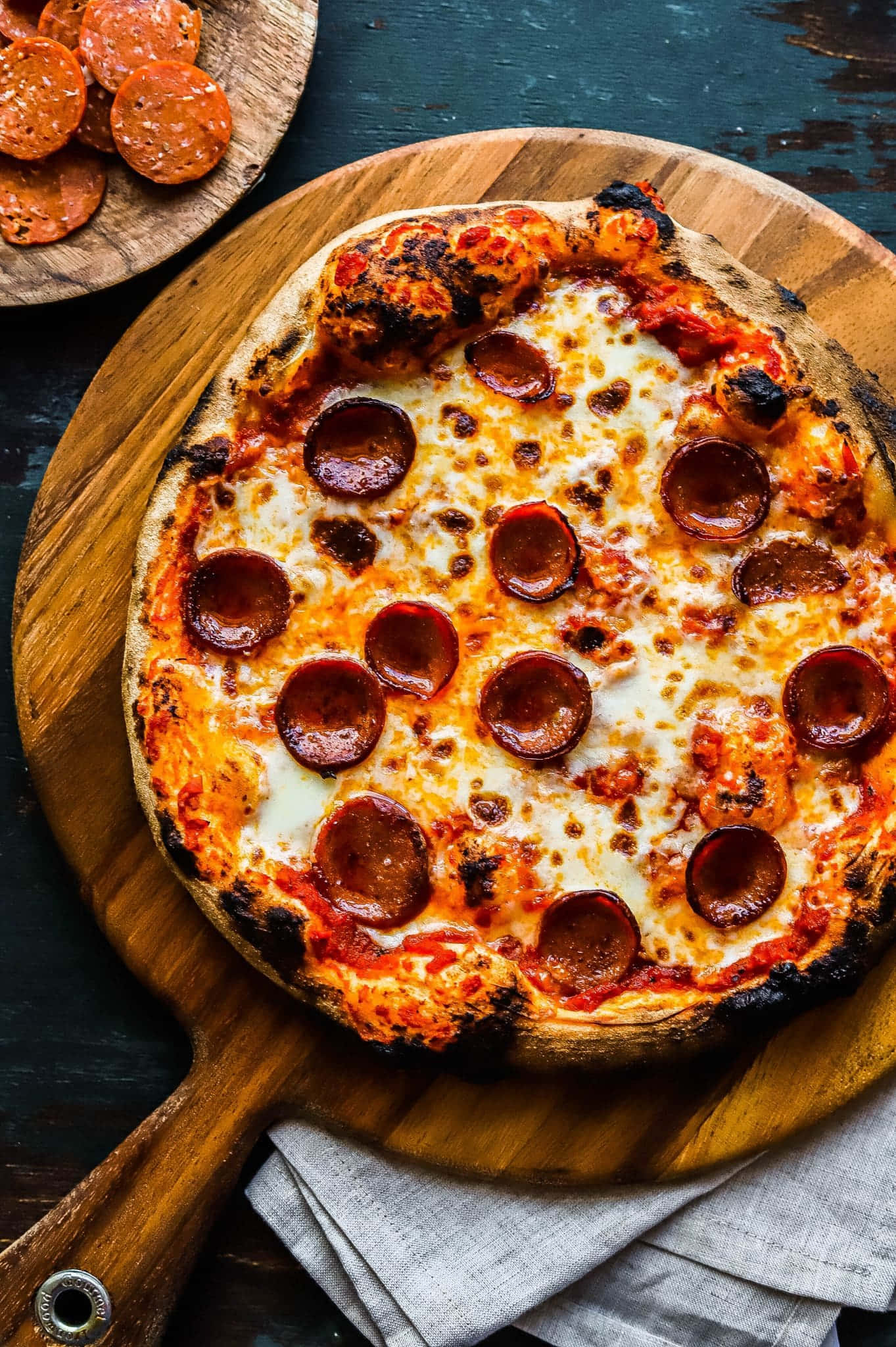 Pizza 桌布 壁紙 — 新鮮出爐的義大利辣香腸披薩，帶有焦脆的餅皮，放在木鏟上，旁邊有一疊餐巾和額外的義大利辣香腸。
