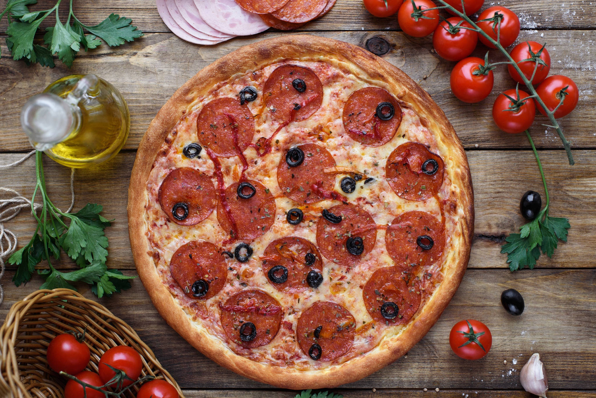 Pizza 桌布 壁紙 — 新鮮出爐的義大利辣香腸披薩配上黑橄欖，放在質樸的木桌上，周圍有櫻桃番茄、歐芹和橄欖油。