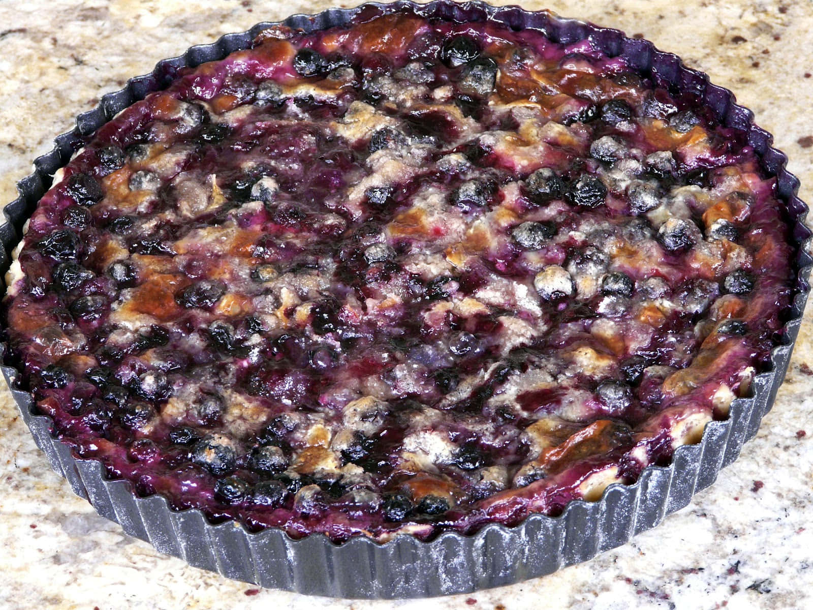 Blueberries Tart 桌布 壁紙 — 新鮮出爐的藍莓塔，多汁的莓果和金黃色的外皮，放在一個有凹槽的金屬烤盤中，置於斑點點的工作檯面上。