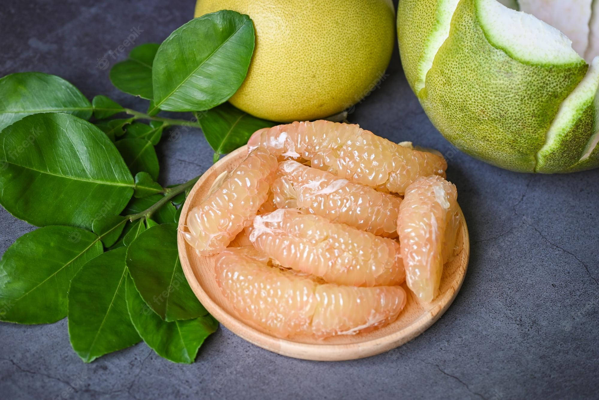 Fruits 桌布 壁紙 — 新鮮剝好的柚子瓣在木碗中閃閃發光，旁邊放著完整的柚子和鮮綠的葉子，置於深色紋理表面上。