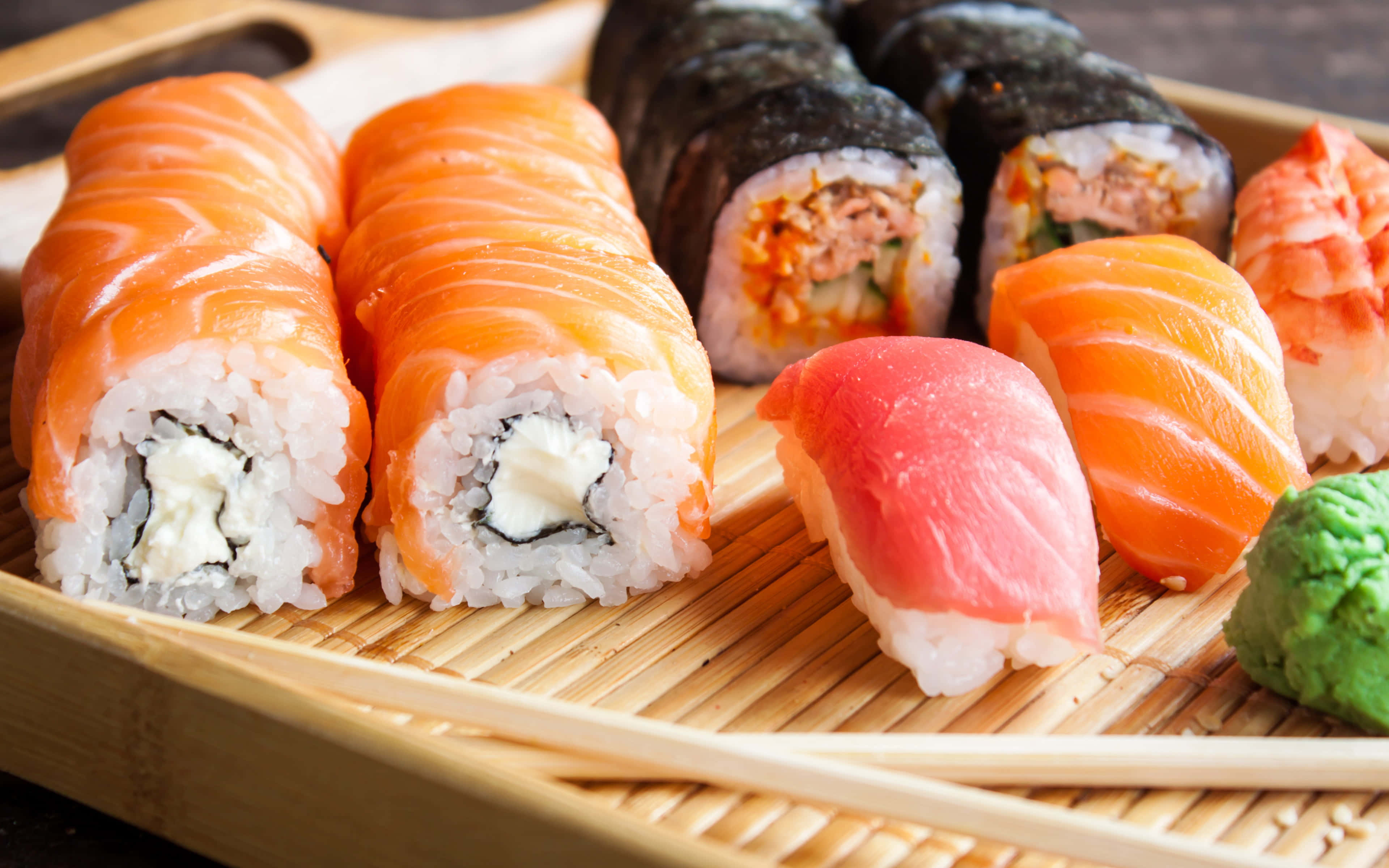 Salmon 桌布 壁紙 — 新鮮壽司捲和握壽司，搭配鮭魚、鮪魚和酪梨，擺放在竹蓆上，附有芥末和筷子。