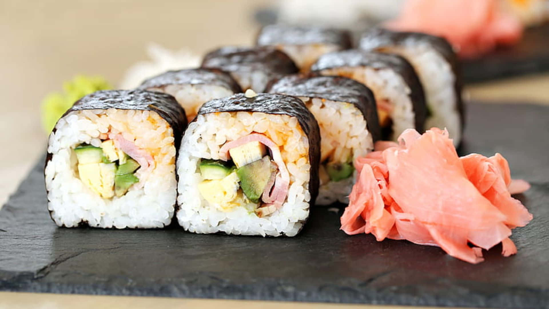 Sushi 桌布 壁紙 — 新鮮壽司捲配酪梨、雞蛋和火腿，整齊地放在石板盤上，旁邊有醃漬生薑。