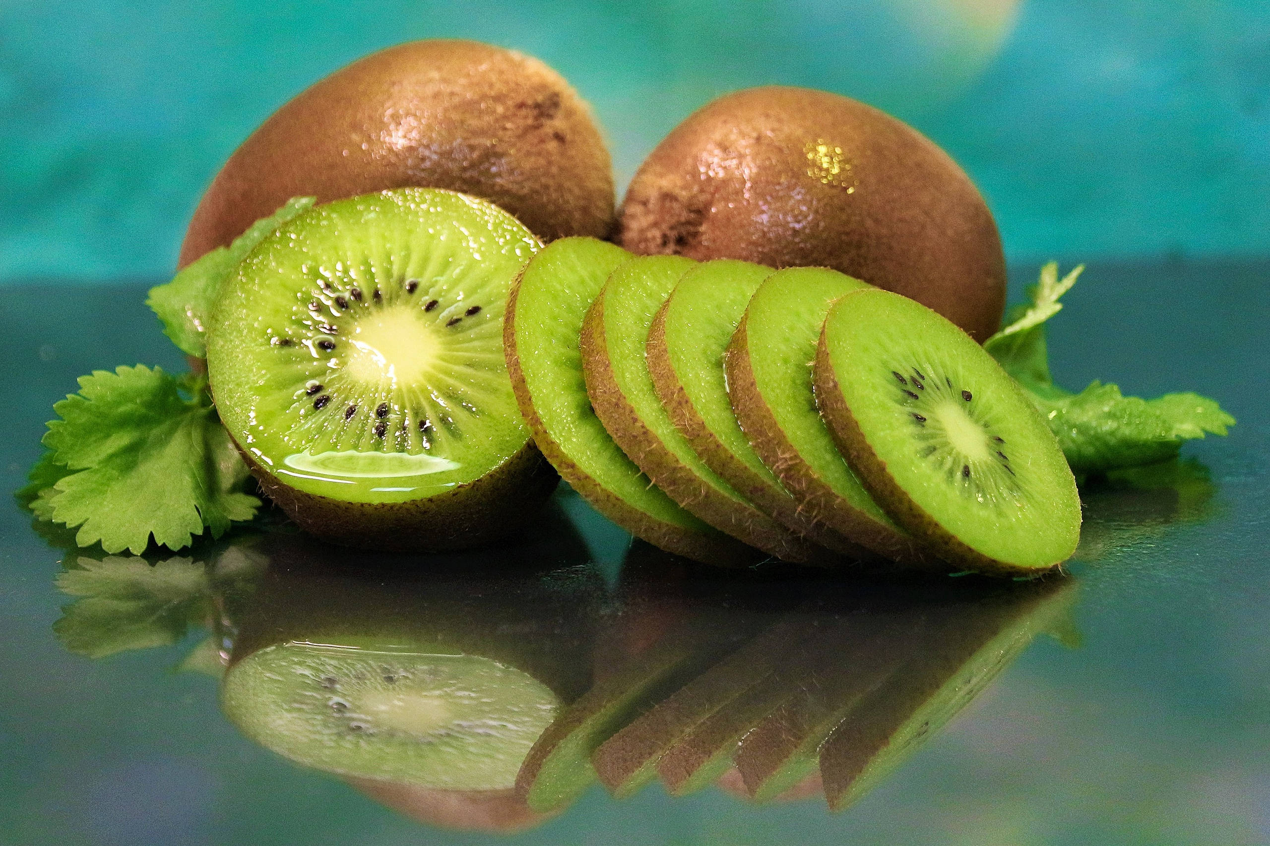 Kiwi 桌布 壁紙 — 新鮮奇異果切片和完整果實，放在光澤感的青綠色表面上，搭配鮮綠葉片和清晰倒影。