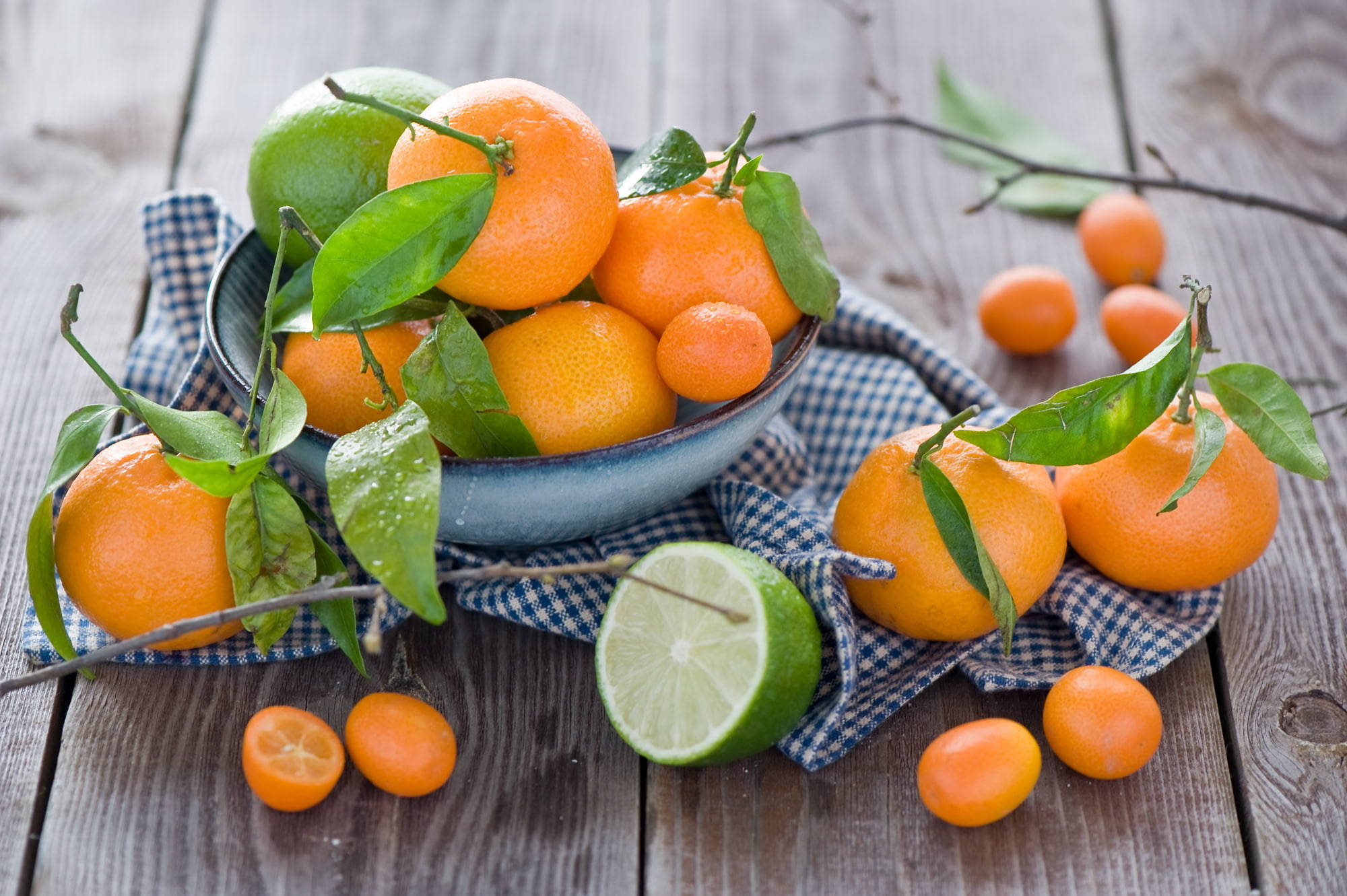 citrus 桌布 壁紙 — 新鮮柑橘類水果，包括柳橙和金桔，連同綠葉從藍色碗中溢出，落在風化木桌上的格紋布上。