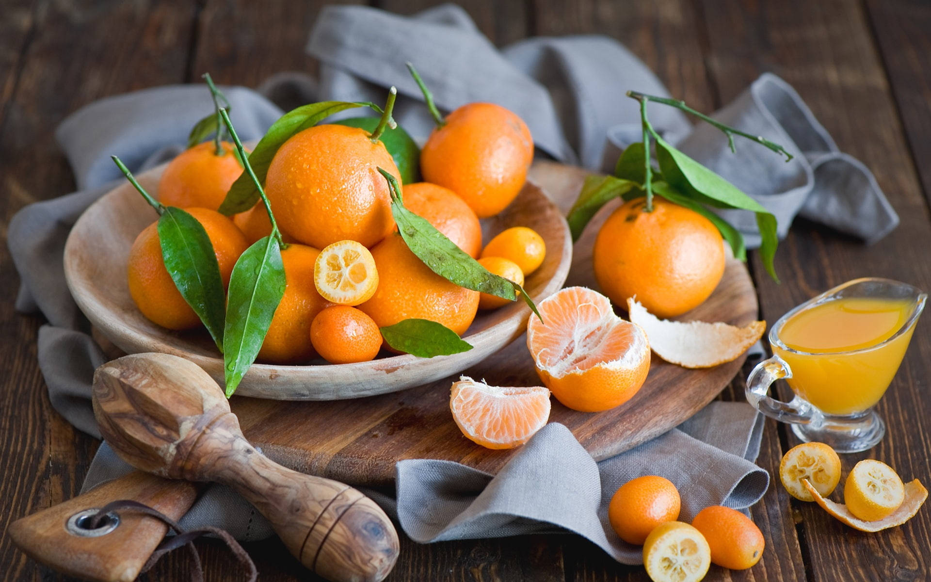 citrus 桌布 壁紙 — 新鮮柑橘類水果連葉放在木碗中，置於鄉村風格桌上，配有一杯果汁和柑橘榨汁器。