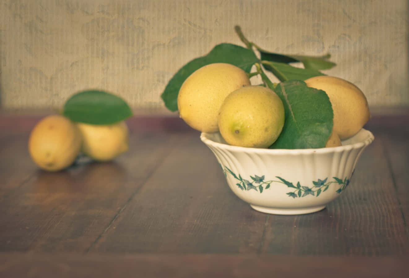 Fruits 桌布 壁紙 — 新鮮檸檬配綠葉，放在白色花卉碗中，置於深色木桌上，背景為有紋理的牆壁。