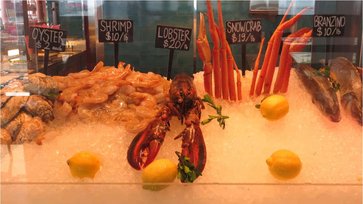 Carnival Conquest 桌布 壁紙 — 新鮮海鮮展示，包括龍蝦、蝦、雪蟹和鱸魚，放在冰上，附有價格牌和檸檬。