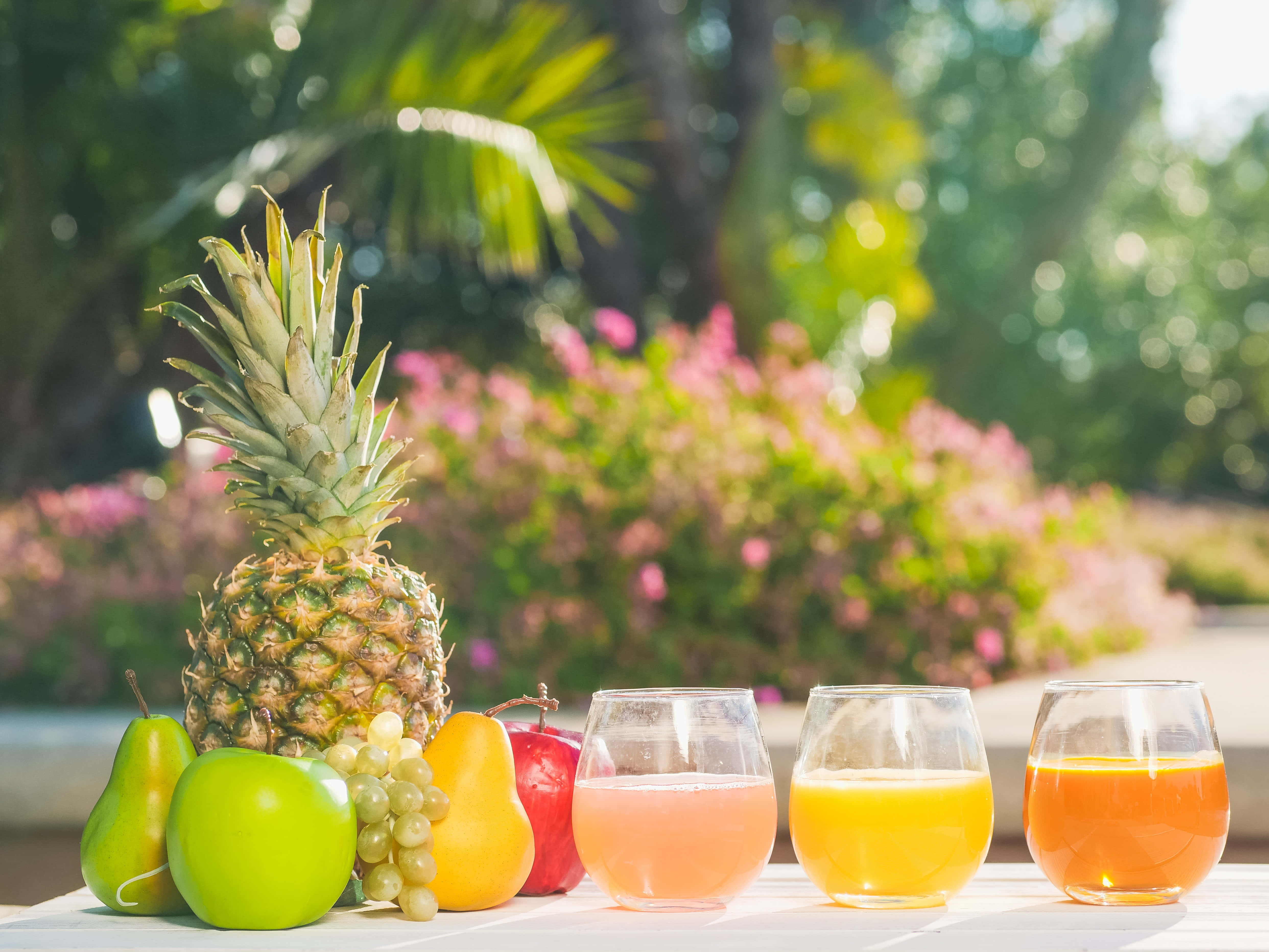 Summer Drinks 桌布 壁紙 — 新鮮熱帶水果和色彩繽紛的果汁在明亮陽光下擺放在桌上，背景有模糊的綠色植物和粉紅色花朵。