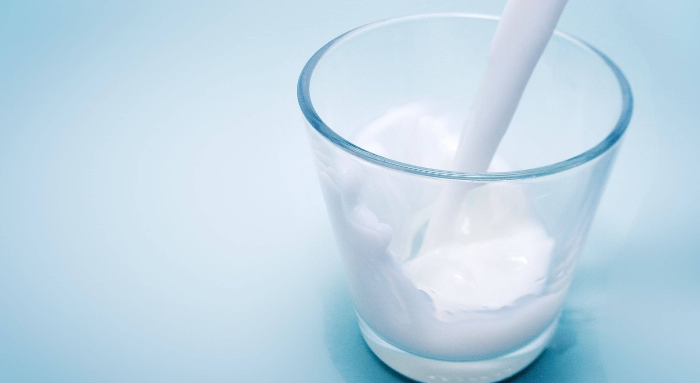 Milk 桌布 壁紙 — 新鮮牛奶倒入透明玻璃杯，以柔和藍色背景襯托，形成輕柔漣漪和奶油漩渦。