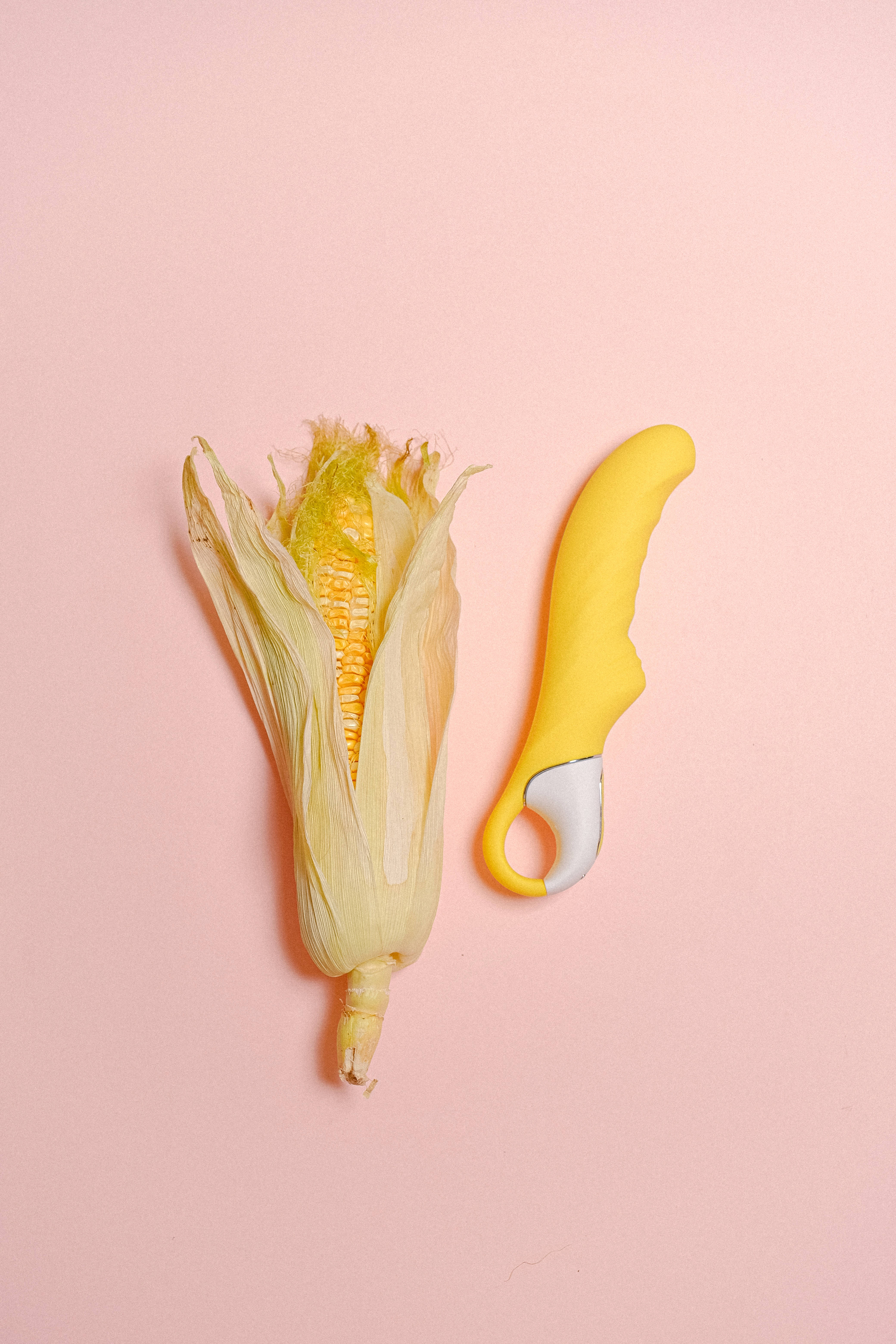Sexual 桌布 壁紙 — 新鮮玉米，部分剝皮，旁邊有一個黃色玉米剝皮器，放在柔和的粉紅色背景上。