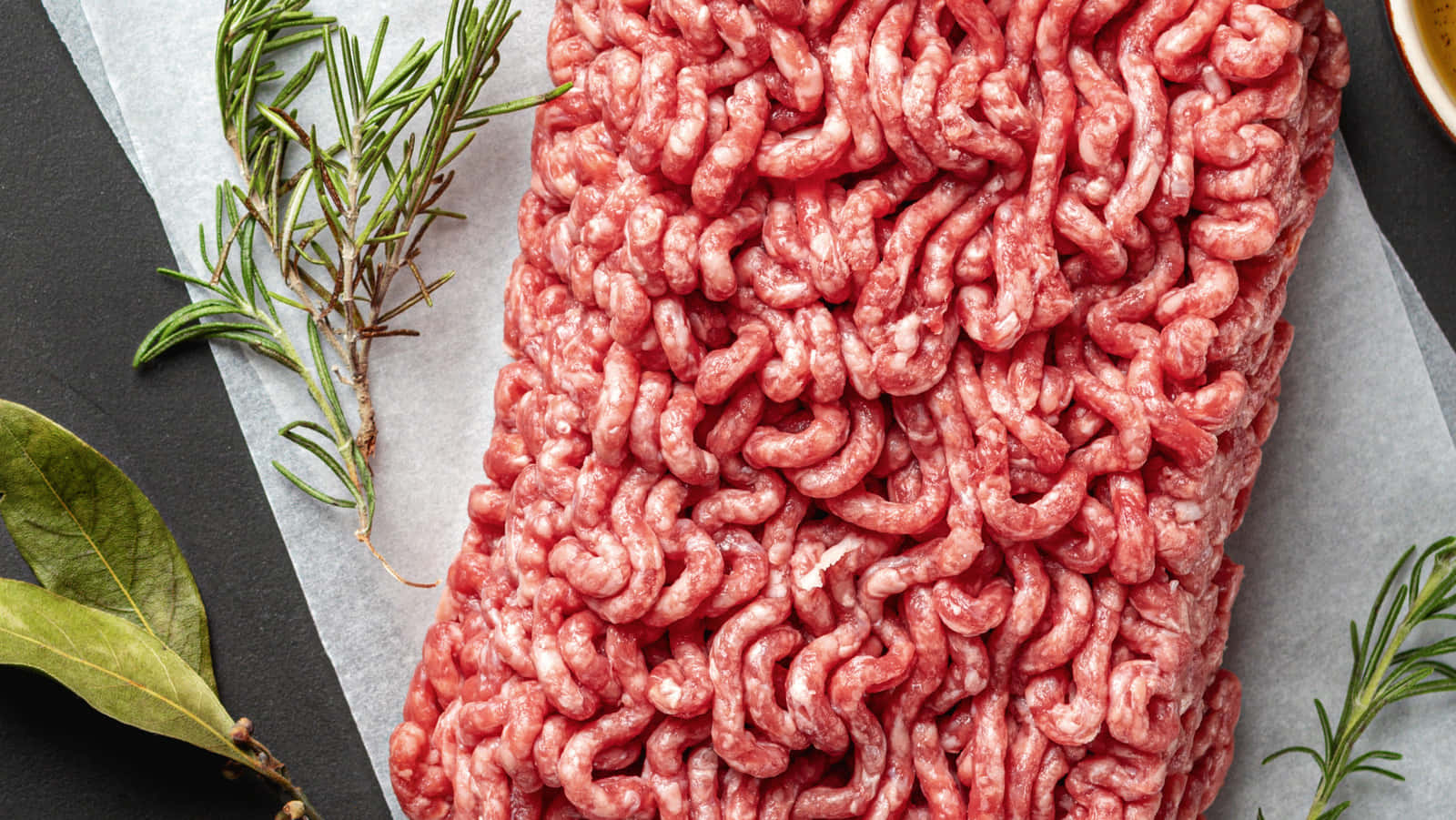 Red Meat 桌布 壁紙 — 新鮮生牛絞肉放在烘焙紙上，旁邊有迷迭香枝和月桂葉，置於深色石板表面上。