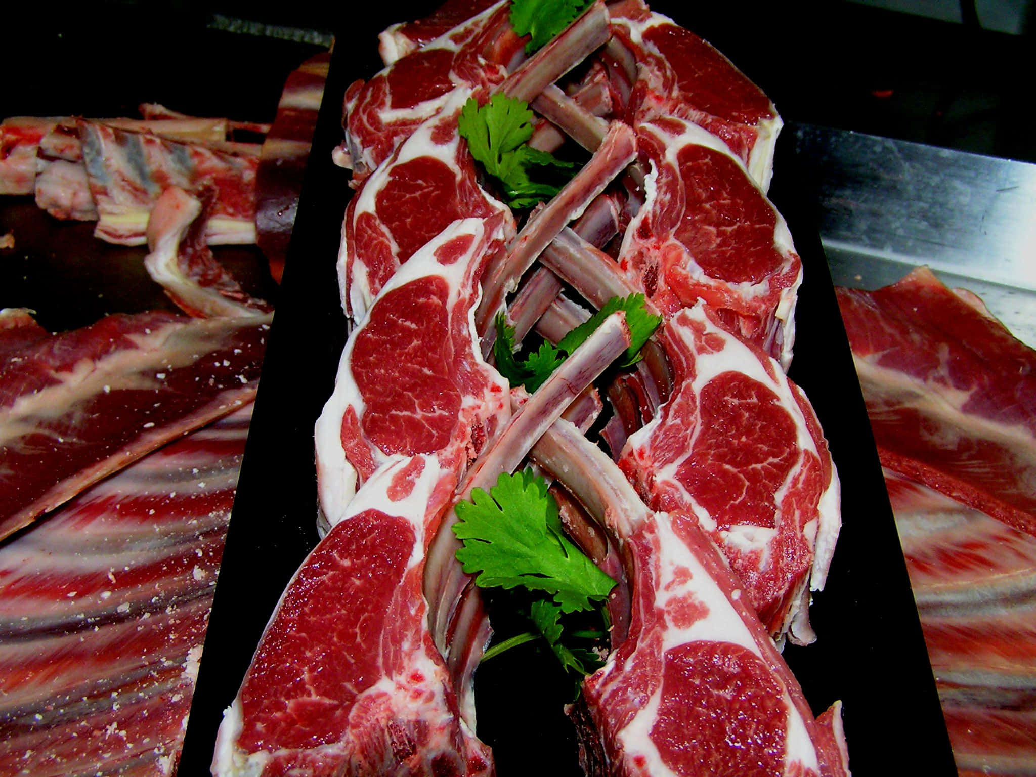 Red Meat 桌布 壁紙 — 新鮮生羊排帶骨，搭配歐芹裝飾，排列在黑色托盤上，背景深色。