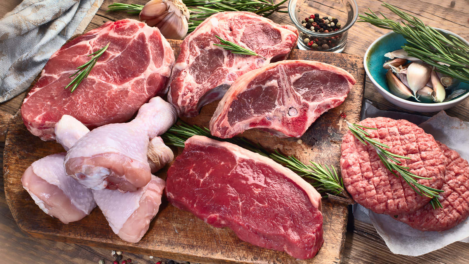 Red Meat 桌布 壁紙 — 新鮮生肉包括牛排、雞大腿和漢堡肉餅，連同香草和大蒜擺放在質樸的木砧板上。