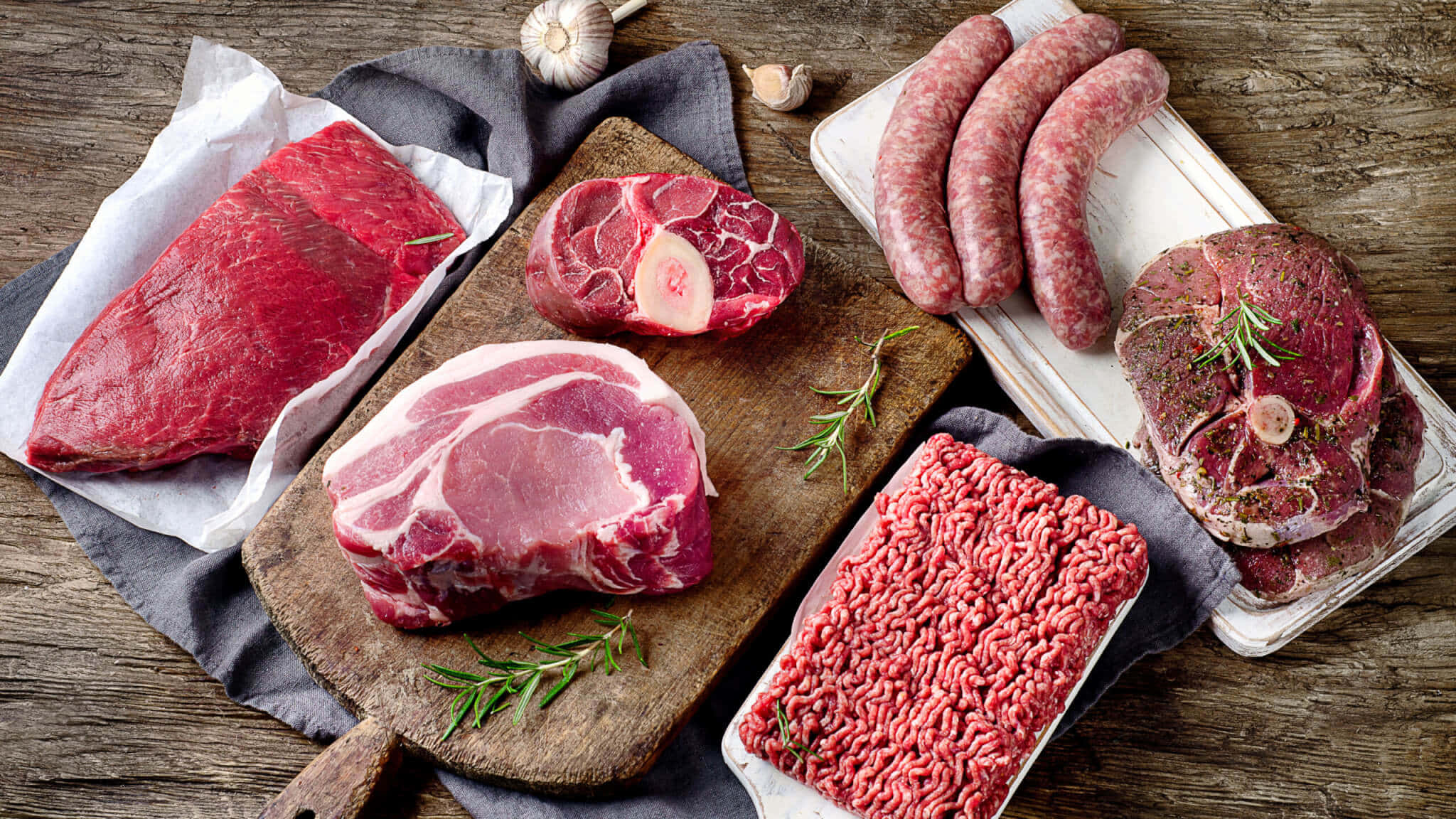 Red Meat 桌布 壁紙 — 新鮮生肉包括牛排、香腸和牛絞肉，放在木板上，搭配迷迭香和大蒜，置於鄉村風木桌上。