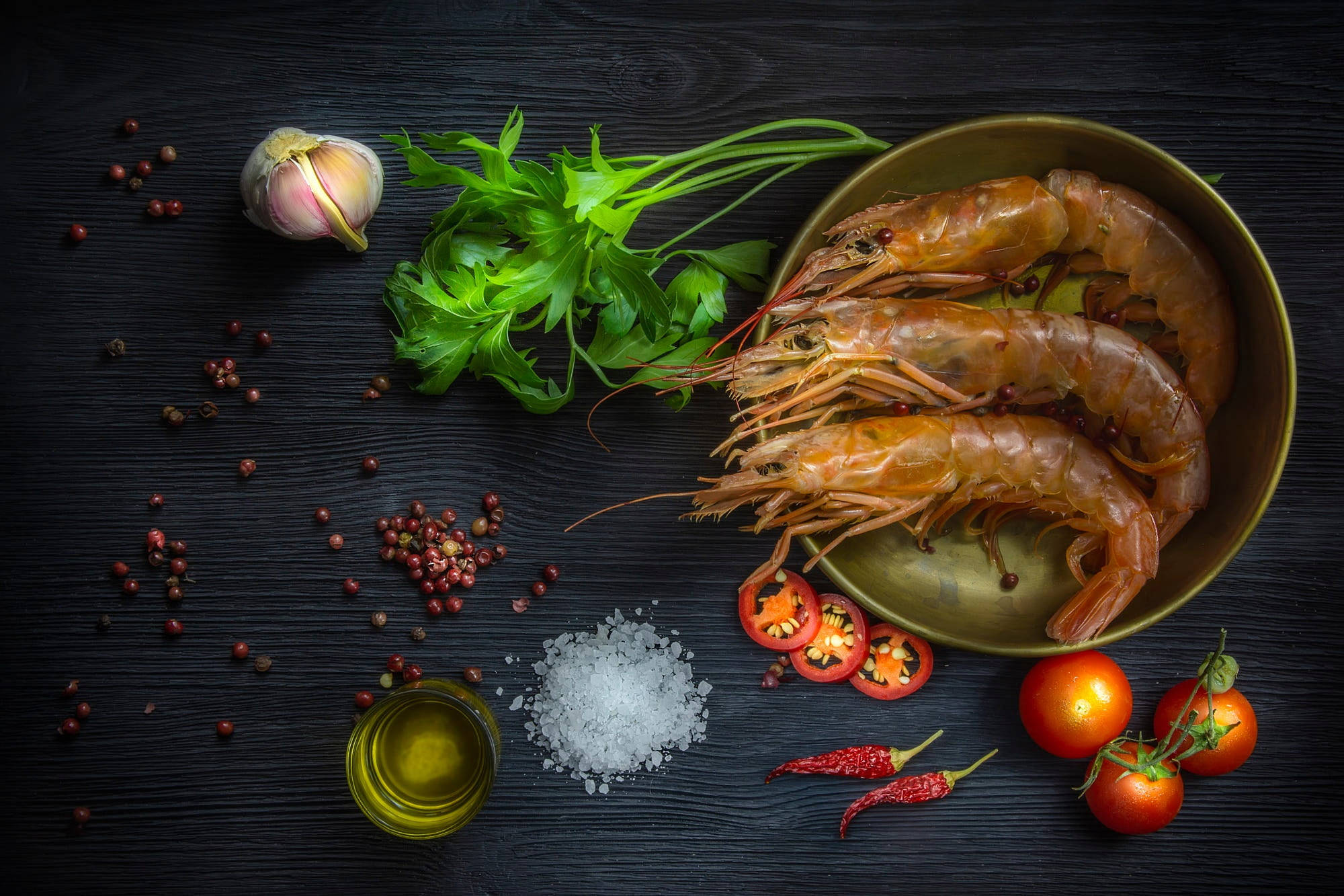 tomato 桌布 壁紙 — 新鮮生蝦放在黃銅碗中，周圍有蒜頭、歐芹、胡椒粒、鹽、油和櫻桃番茄，置於深色木質表面上。