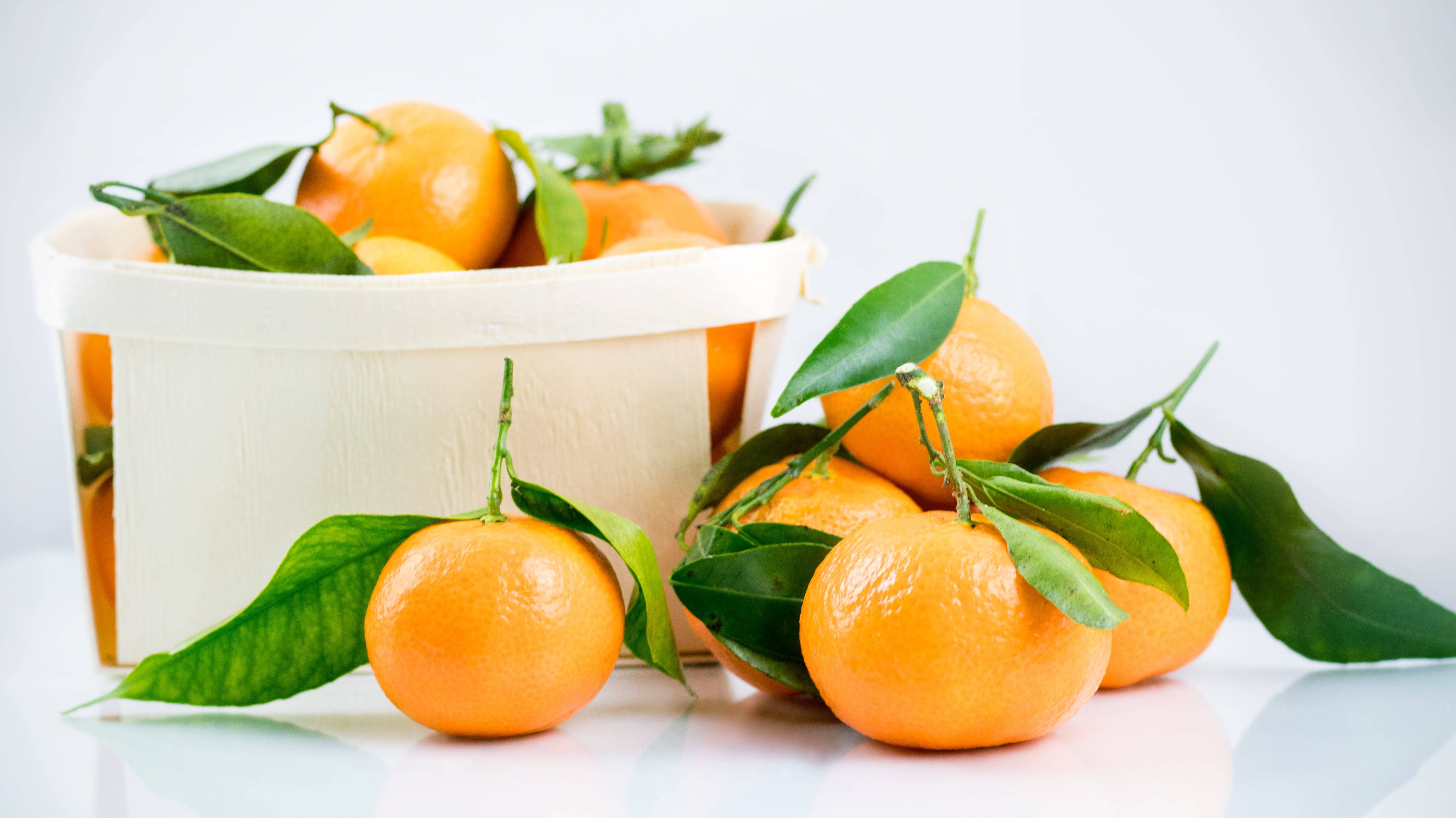 Fruits 桌布 壁紙 — 新鮮的柑橘連同綠葉從白色籃子中溢出，落在反射表面上，背景為明亮的白色。