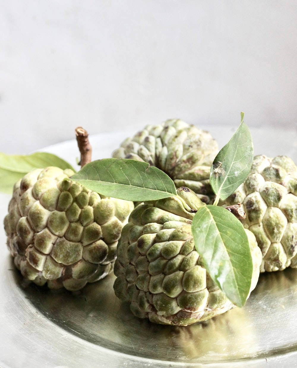 Fruit 桌布 壁紙 — 新鮮的綠色牛油果（custard apples）連葉子放在反射性銀盤上，背景是柔和白色。