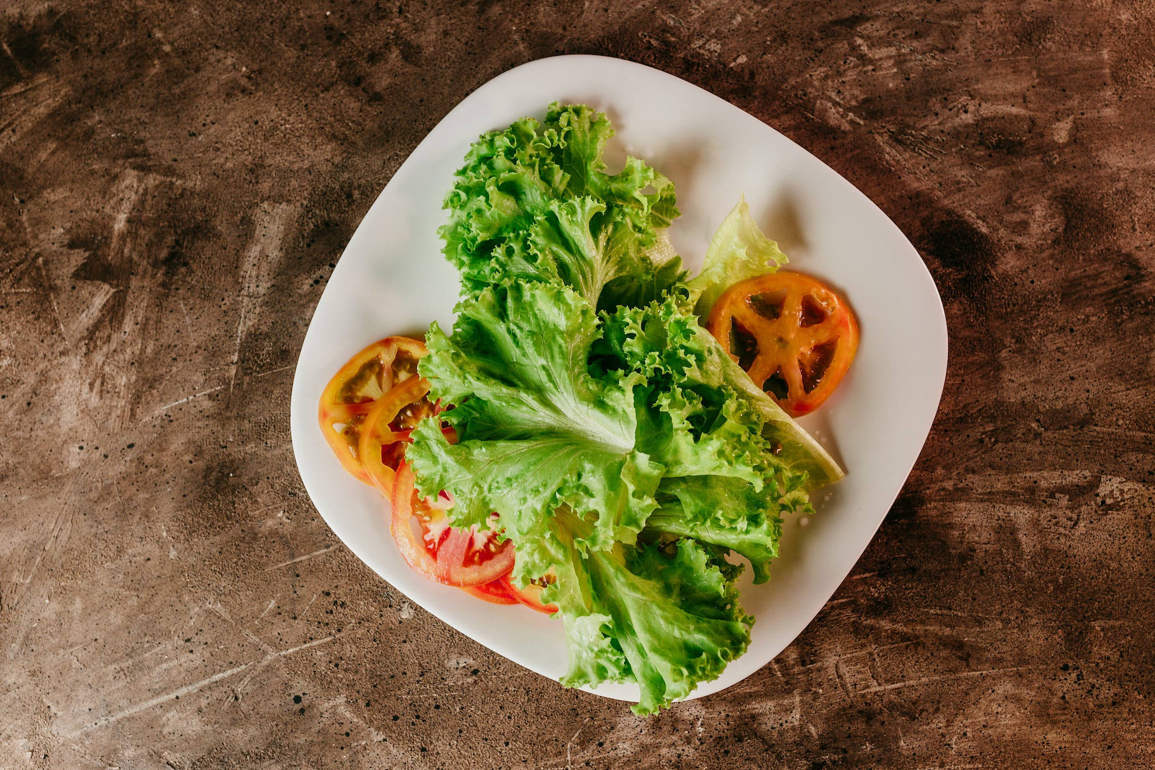 Vegetables 桌布 壁紙 — 新鮮的綠色生菜沙拉搭配切片番茄，放在白色盤子上，置於鄉村風格的棕色紋理表面。