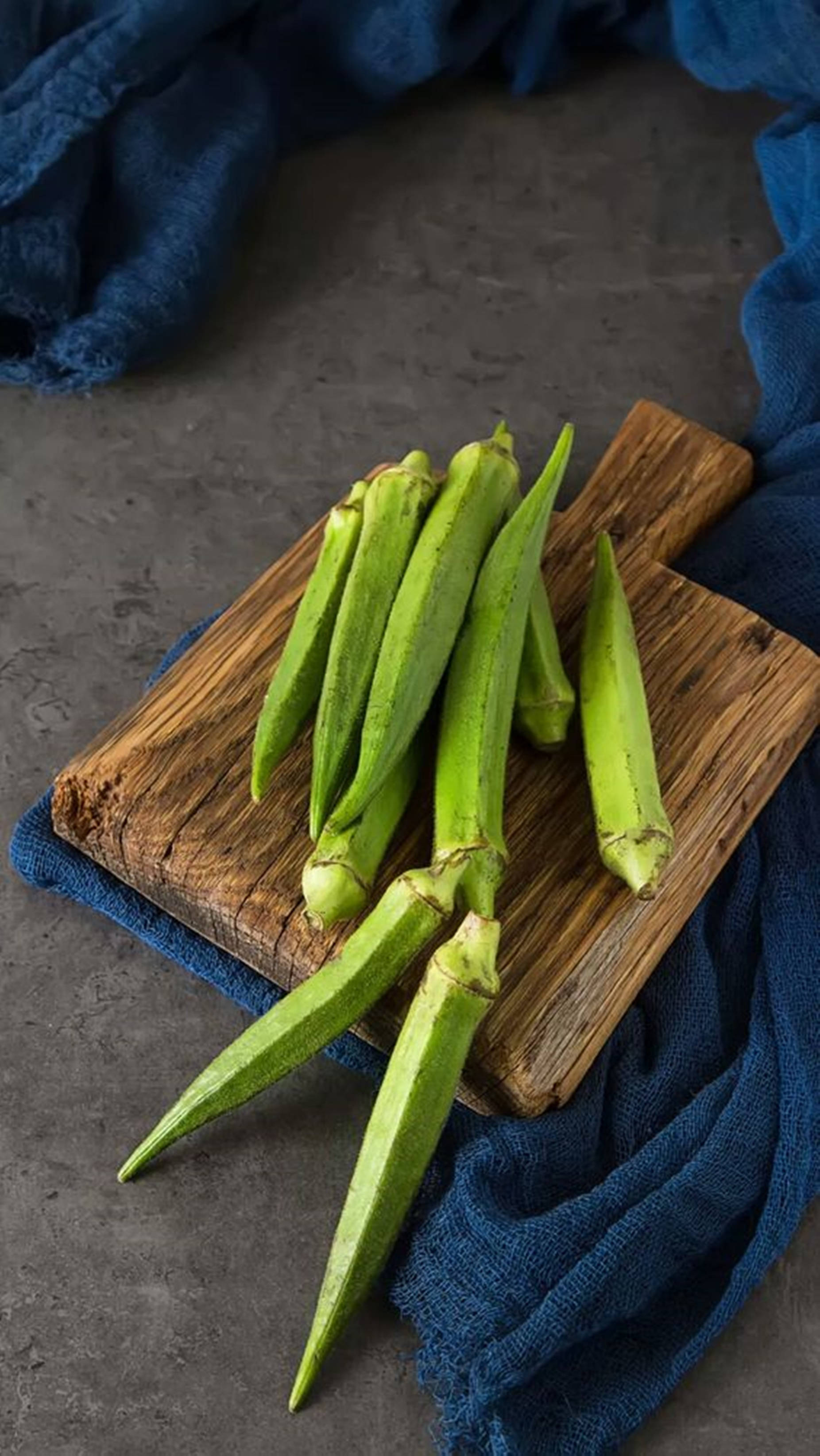 food iPhone 桌布 壁紙 — 新鮮的綠色秋葵莢在灰色石頭背景下，放在鋪了深藍色布料的質樸木砧板上。