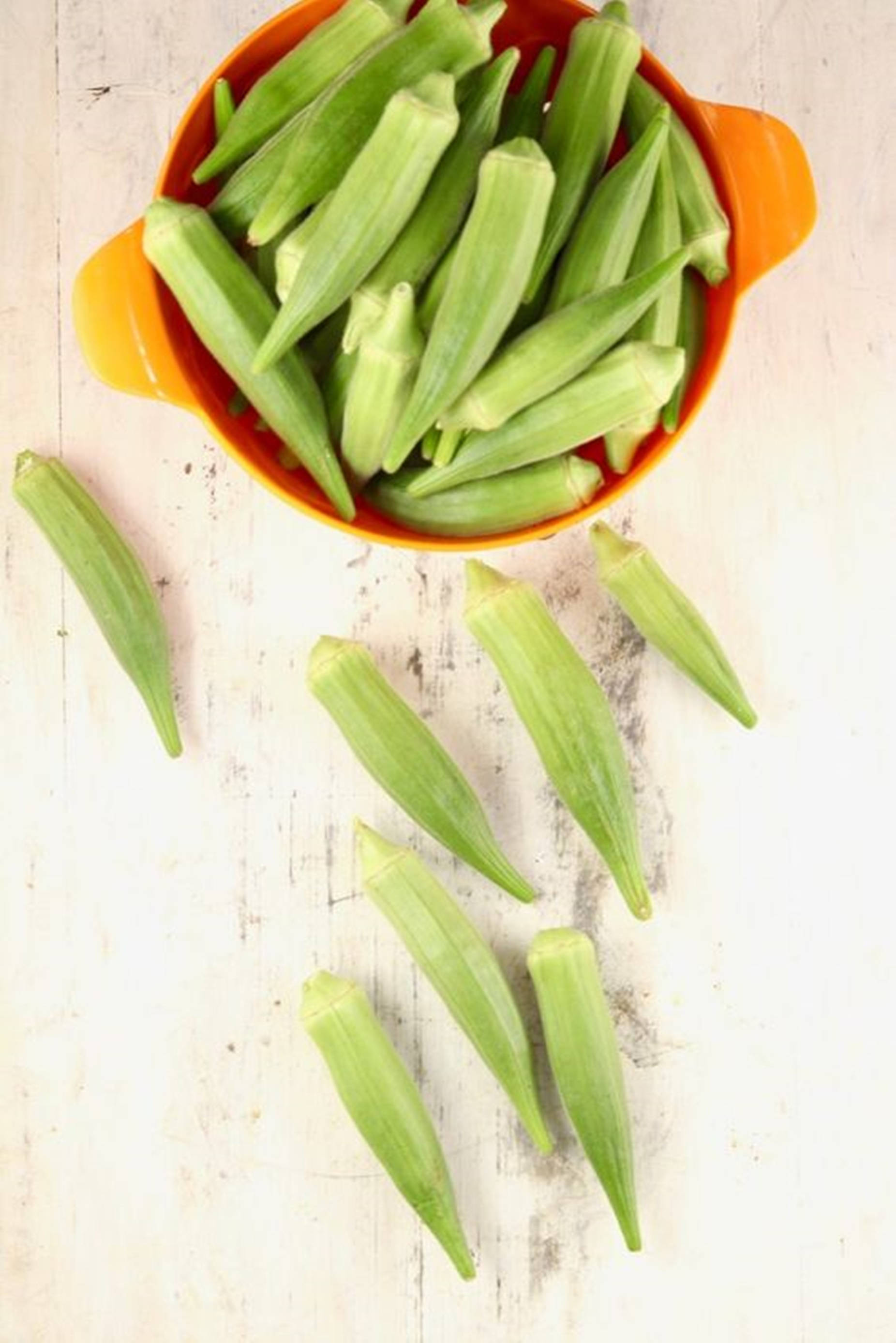 Healthy Food 桌布 壁紙 — 新鮮的綠色秋葵莢從一個亮橙色的碗中溢出，落在一個風化的白色木質表面上。