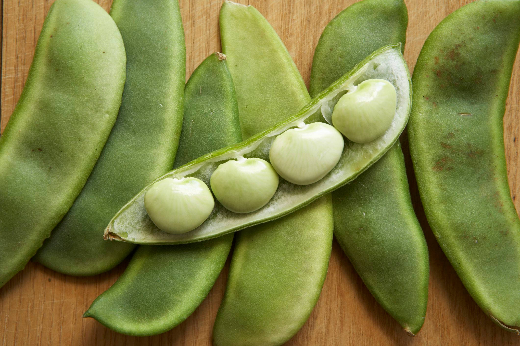 beans 桌布 壁紙 — 新鮮的綠色蠶豆莢，內有飽滿的淡色種子，放在溫暖的木質表面上。