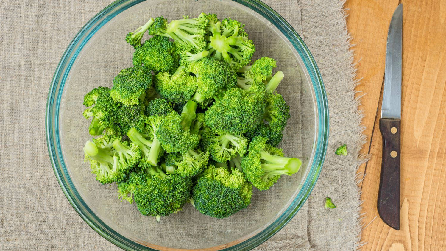花椰菜 桌布 壁紙 — 新鮮的綠色西蘭花小朵填滿一個透明玻璃碗，放在有紋理的布上，旁邊有木桌和一把刀。