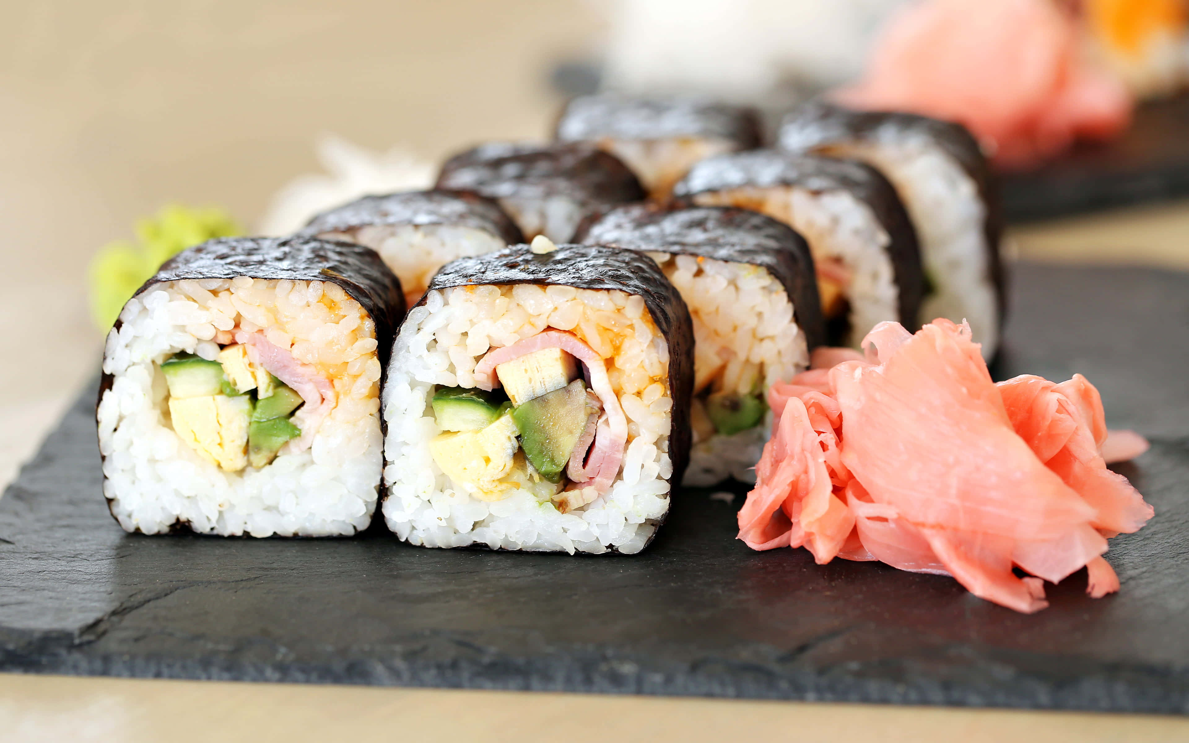 Types Of Sushi 桌布 壁紙 — 新鮮的酪梨火腿壽司卷整齊地放在深色石板盤上，旁邊有鮮豔的醃薑。