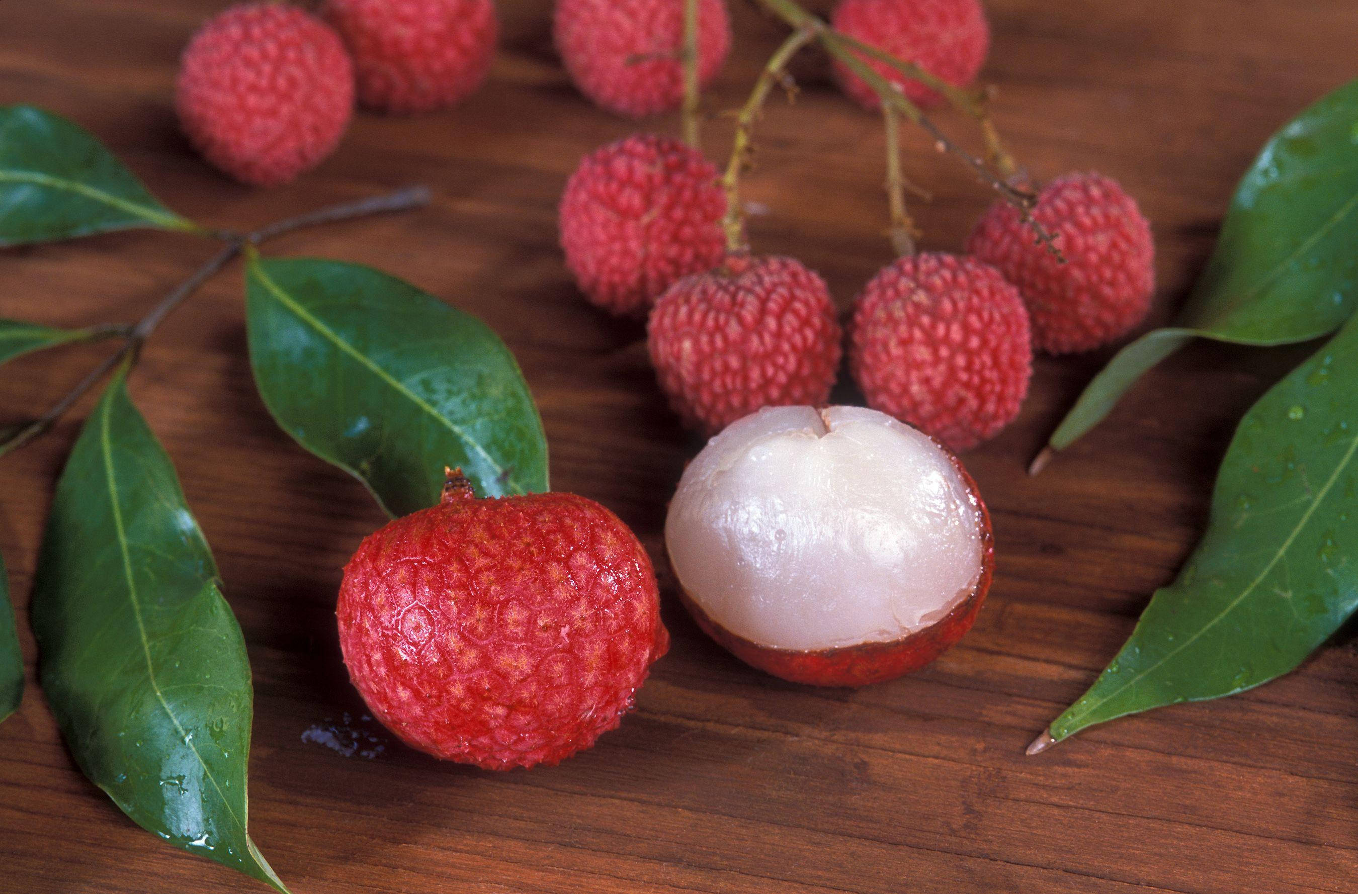 Litchi 桌布 壁紙 — 新鮮紅色荔枝配綠葉放在木質表面上，其中一個裂開露出半透明白色果肉。