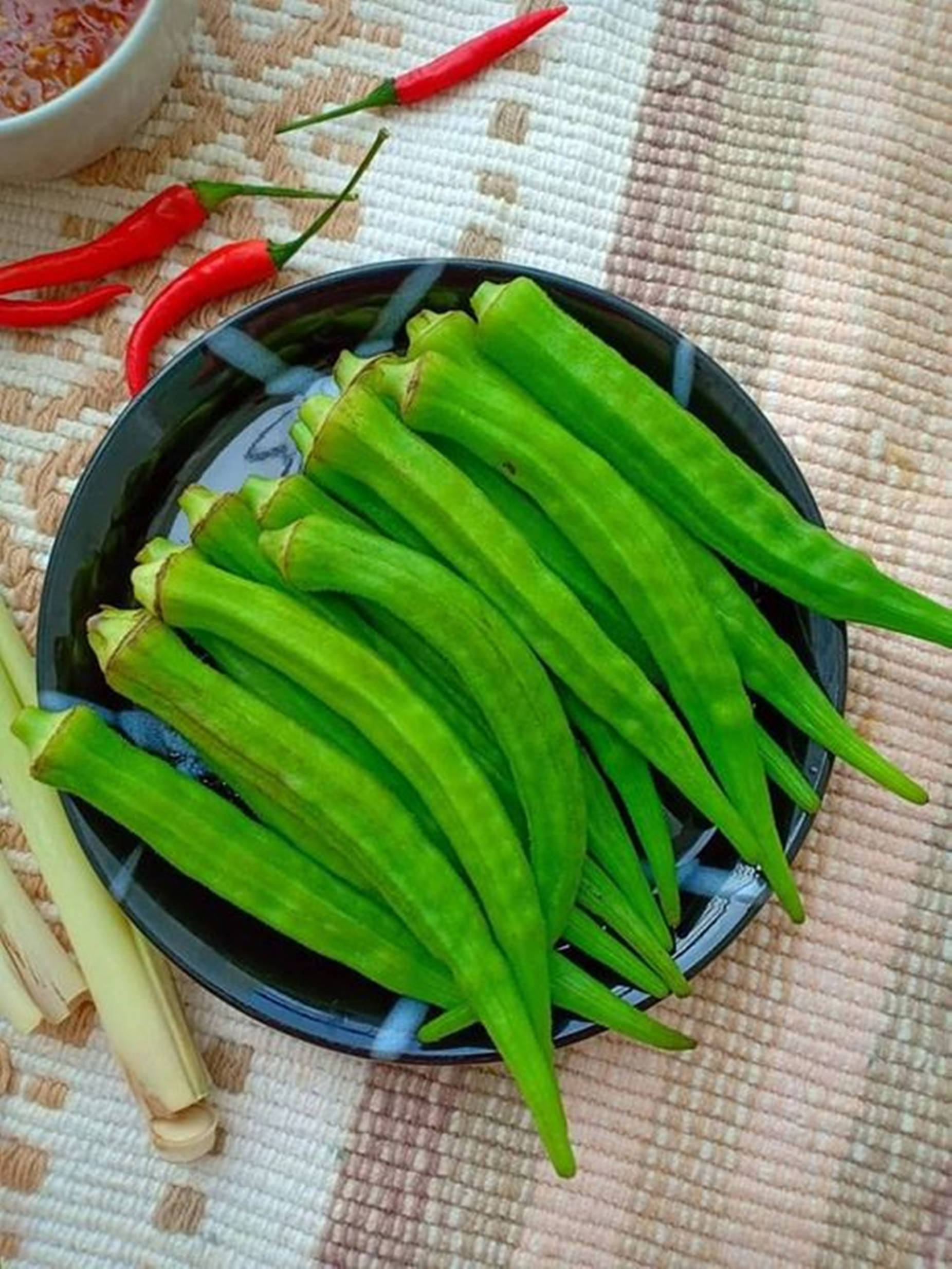 healthy 桌布 壁紙 — 新鮮綠色秋葵莢堆疊在黑色碗中，旁邊有紅辣椒和香茅，放在編織墊上。