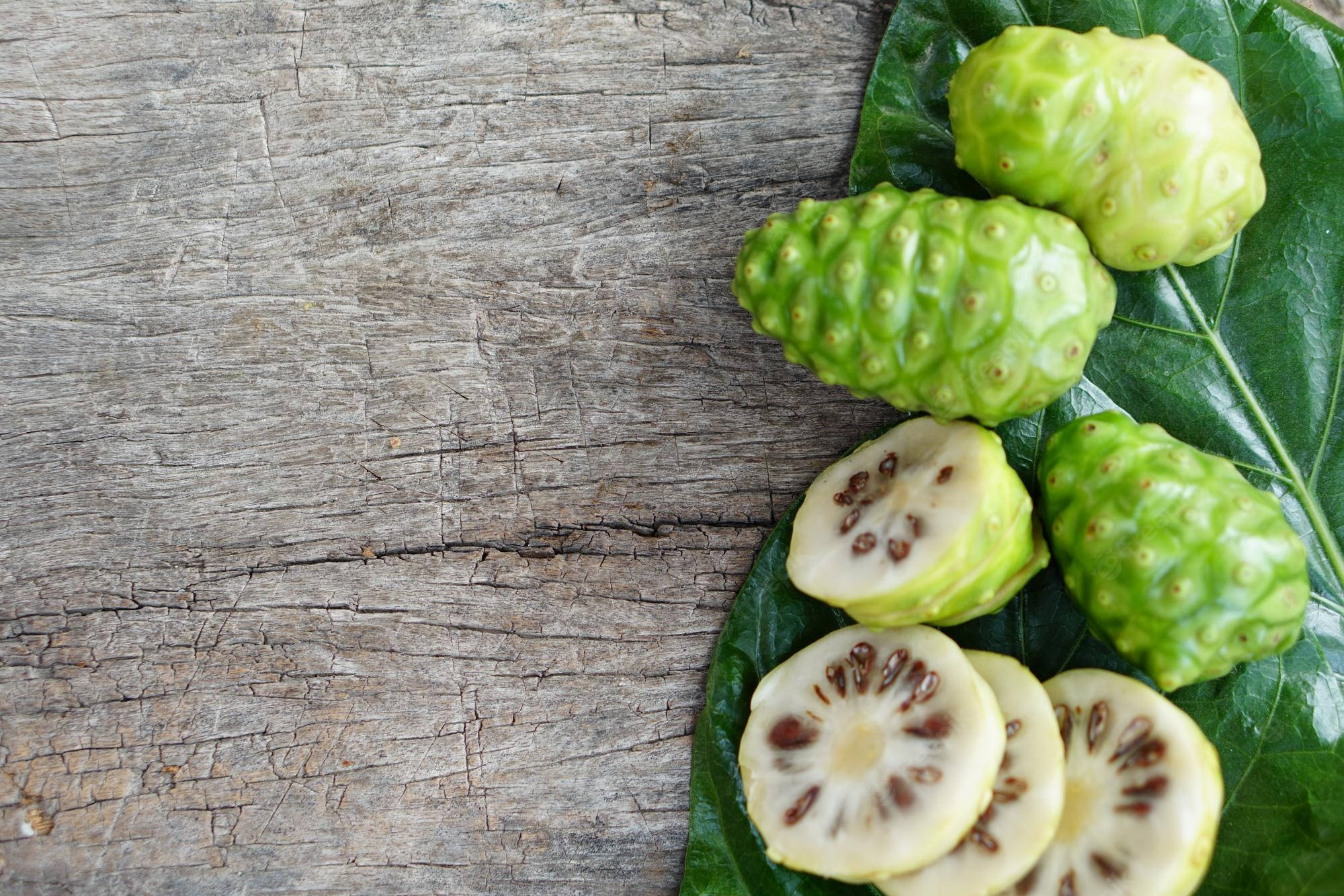 Fruits 桌布 壁紙 — 新鮮綠色諾麗果（完整及切片露出種子）放在大葉上，置於經風化的木質表面。