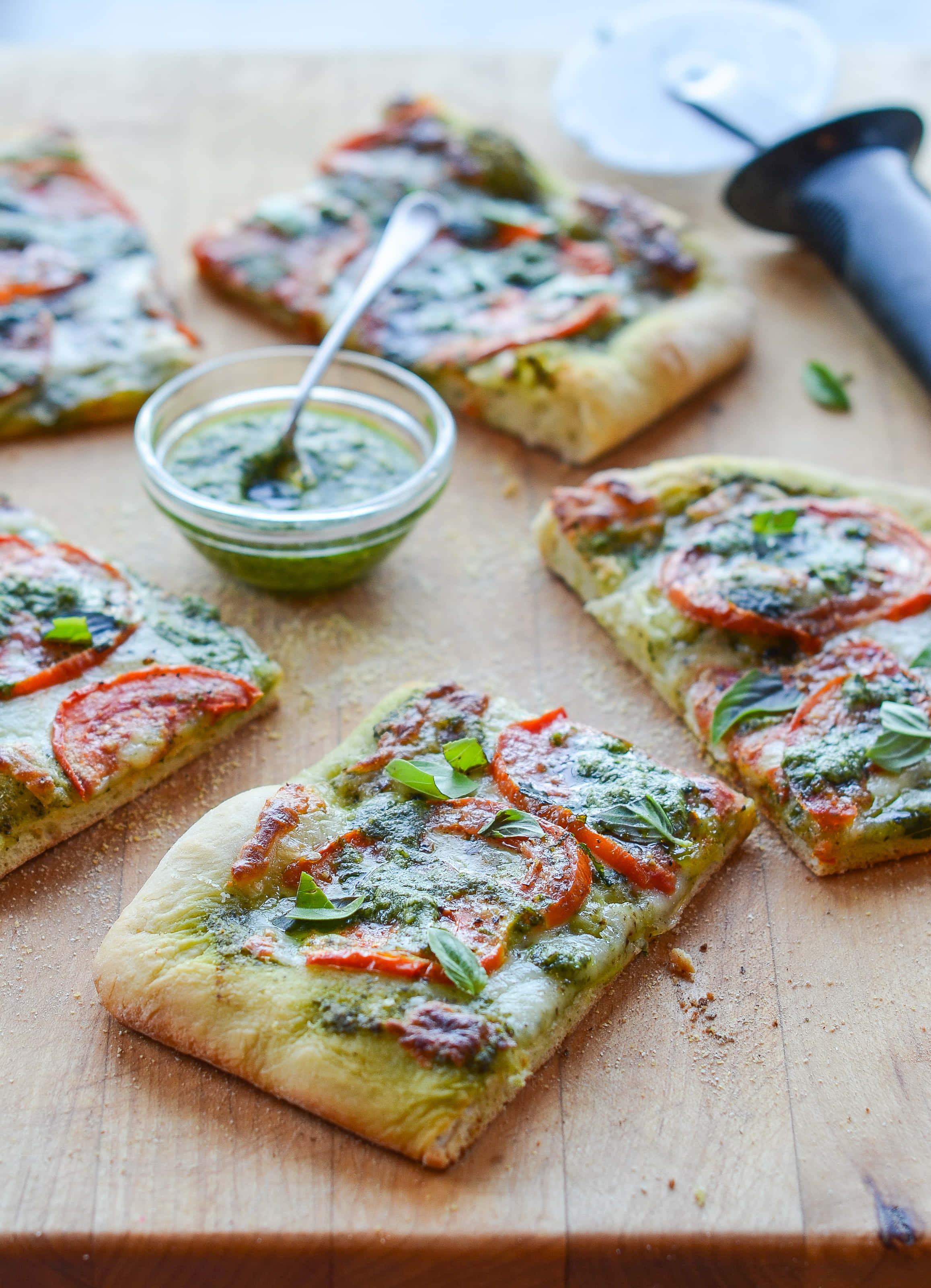 Pizza 桌布 壁紙 — 新鮮羅勒青醬披薩片配番茄和起司放在木板上，旁邊有一小碗青醬和披薩切刀。